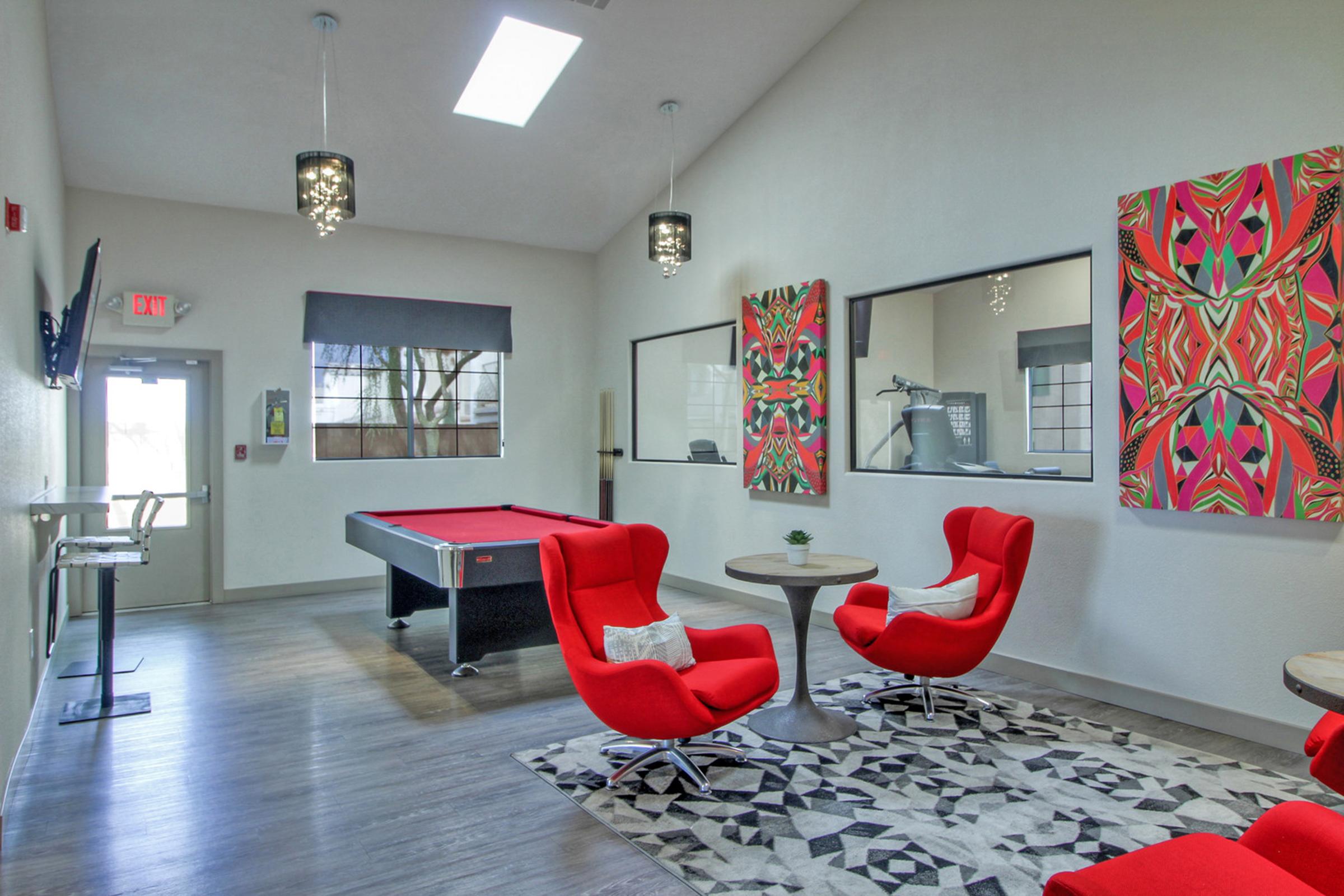Interior of a recreational lounge featuring a red pool table, modern red chairs around a small table, and vibrant abstract artwork on the walls. Large windows allow natural light, and there is a stylish rug on the floor. The space is designed for relaxation and socializing.