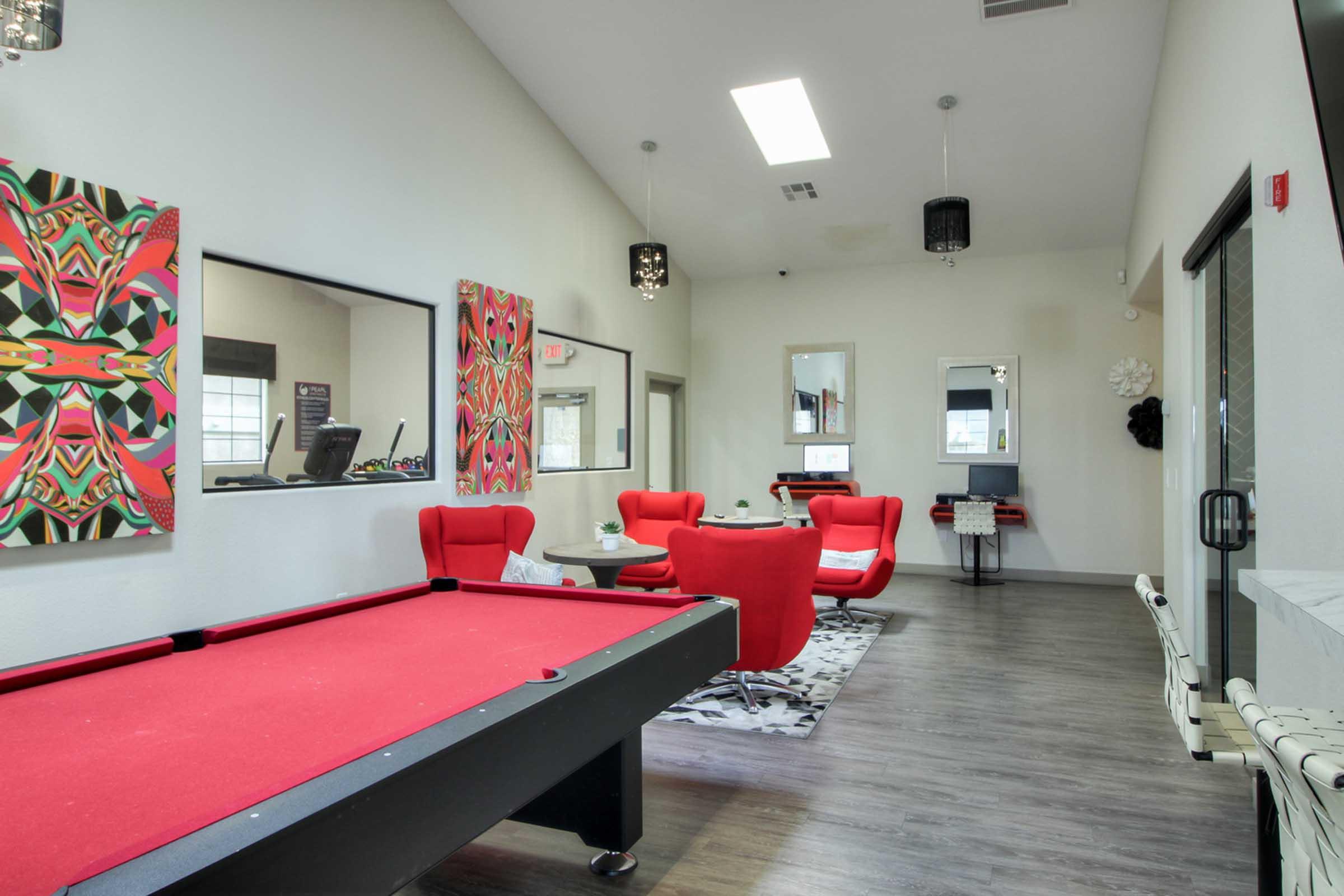 Interior of a modern recreational room featuring a red pool table, red lounge chairs, and a stylish patterned rug. The walls are decorated with colorful artwork, and there are mirrors and lighting fixtures enhancing the space's atmosphere.