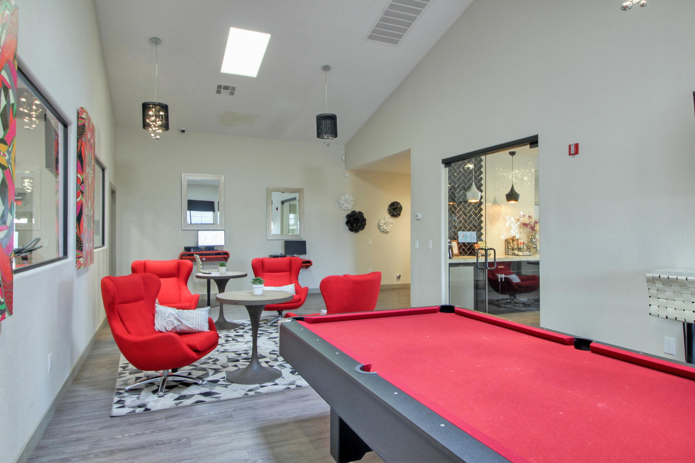 Interior of a modern recreational room featuring vibrant red lounge chairs, a round table, and a pool table with a red felt surface. The space is well-lit with decorative lighting, including pendant lamps, and has large windows. A stylish black and white rug complements the decor, creating a welcoming atmosphere.