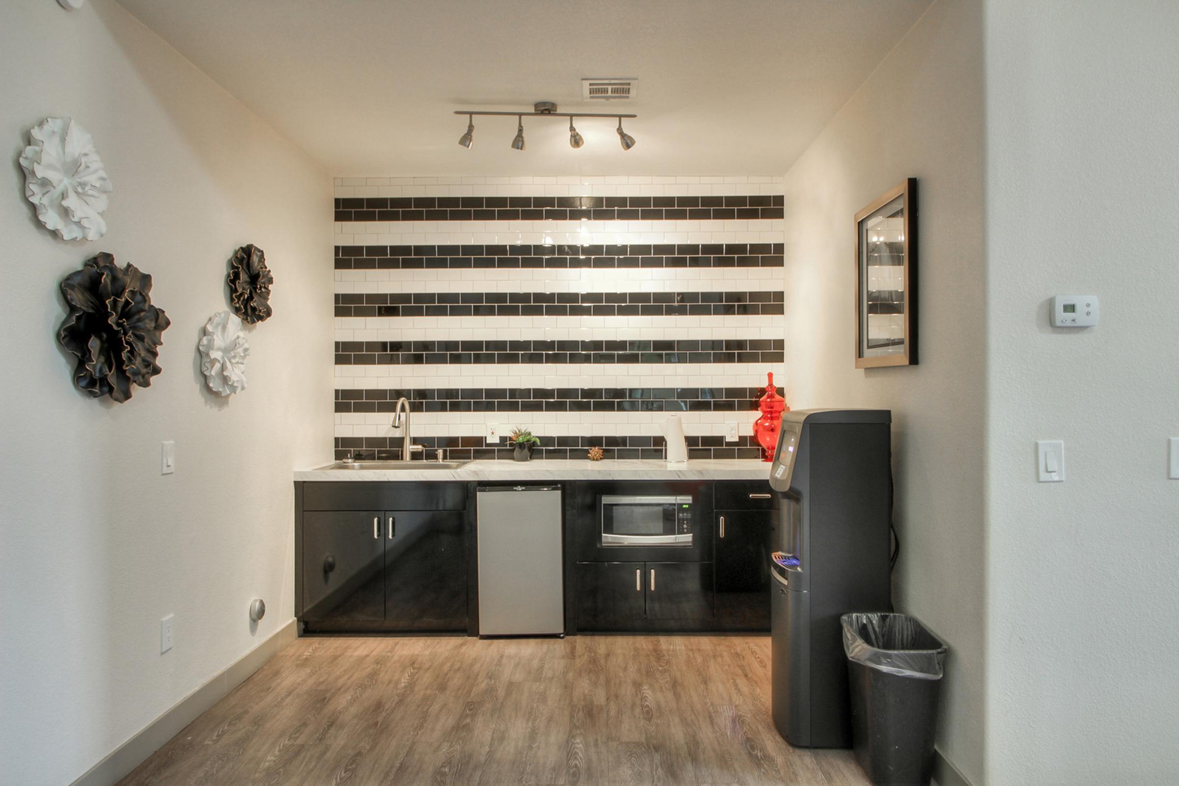Modern kitchen featuring black and white striped tiles as a backsplash, dark cabinetry, a stainless steel sink, microwave, and a small plant. Decorative wall accents include floral wall art. A trash can is placed in the corner, and the floor is a warm wooden design.