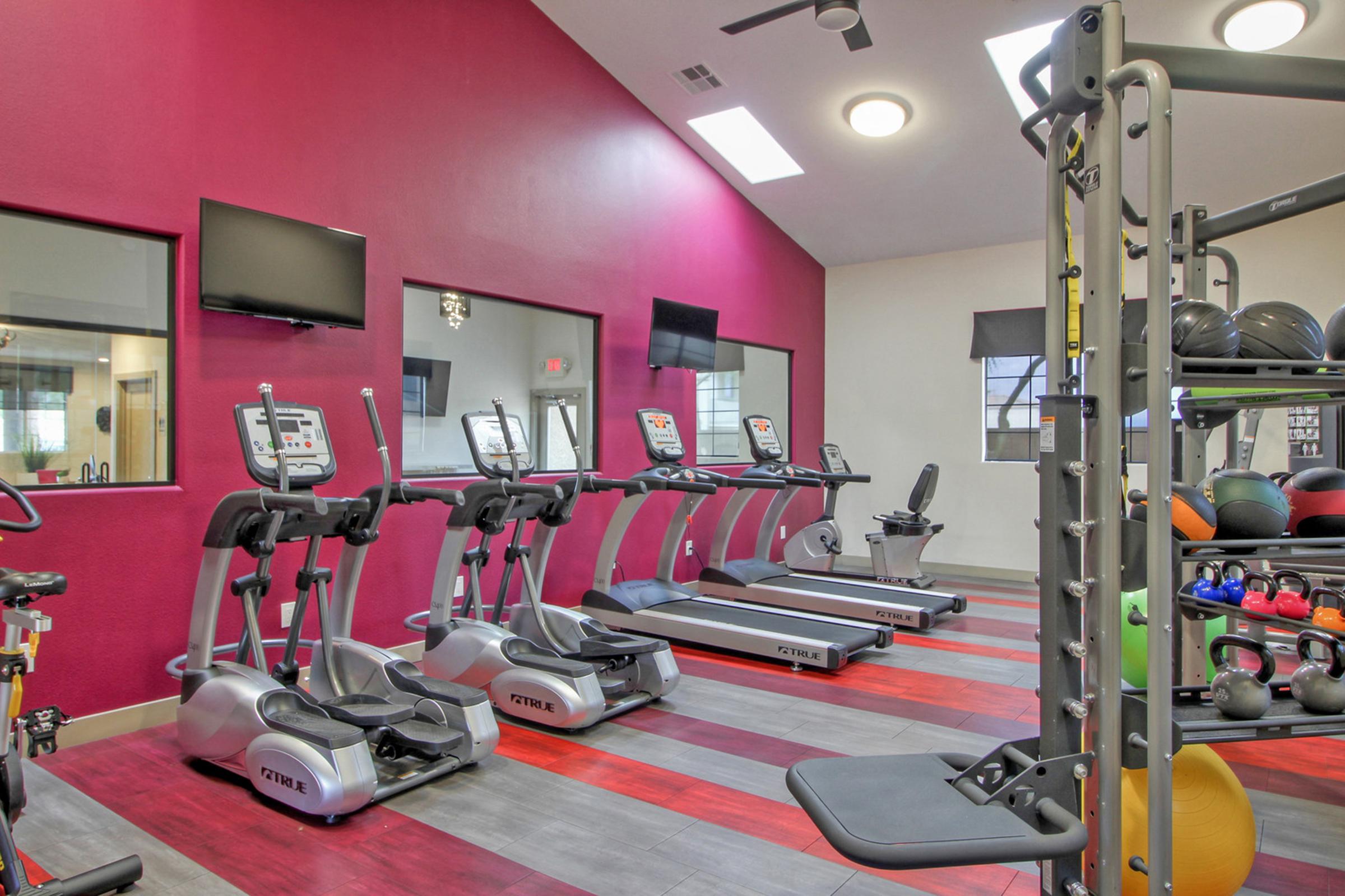 A modern gym interior featuring multiple cardio machines like elliptical trainers and treadmills. The wall is painted in a vibrant pink, and there are large windows with natural light. The space is well-organized with workout equipment, including dumbbells and exercise balls, creating a motivating fitness environment.