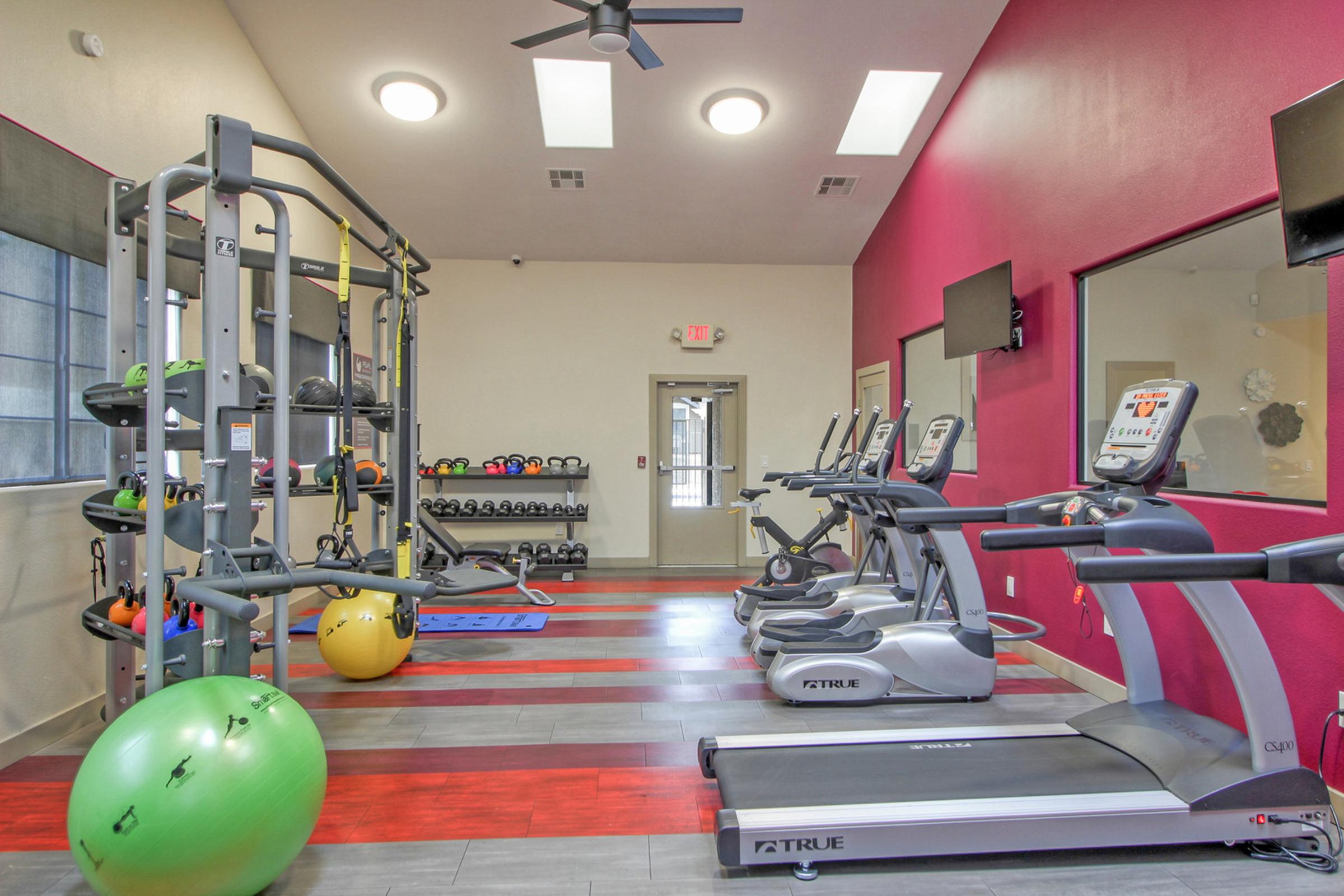 A bright and modern gym featuring exercise equipment including treadmills, ellipticals, and weight machines. Colorful exercise balls and a rack of weights are visible along the wall. The room has large windows allowing natural light and is decorated with red and grey walls.