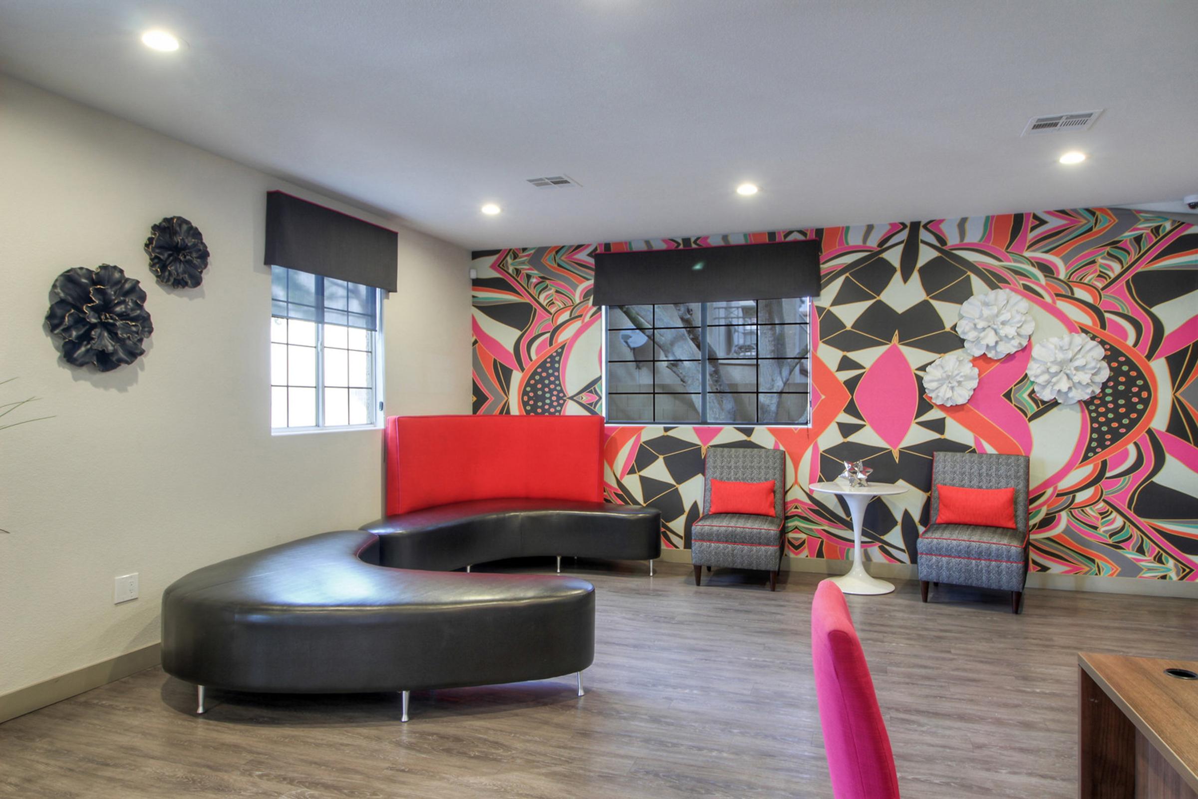A modern lounge area featuring a curved black sofa with red accents, two patterned armchairs, and a small white table. The walls display a vibrant pink and black geometric mural, complemented by decorative wall pieces. Bright lighting enhances the stylish and inviting atmosphere.