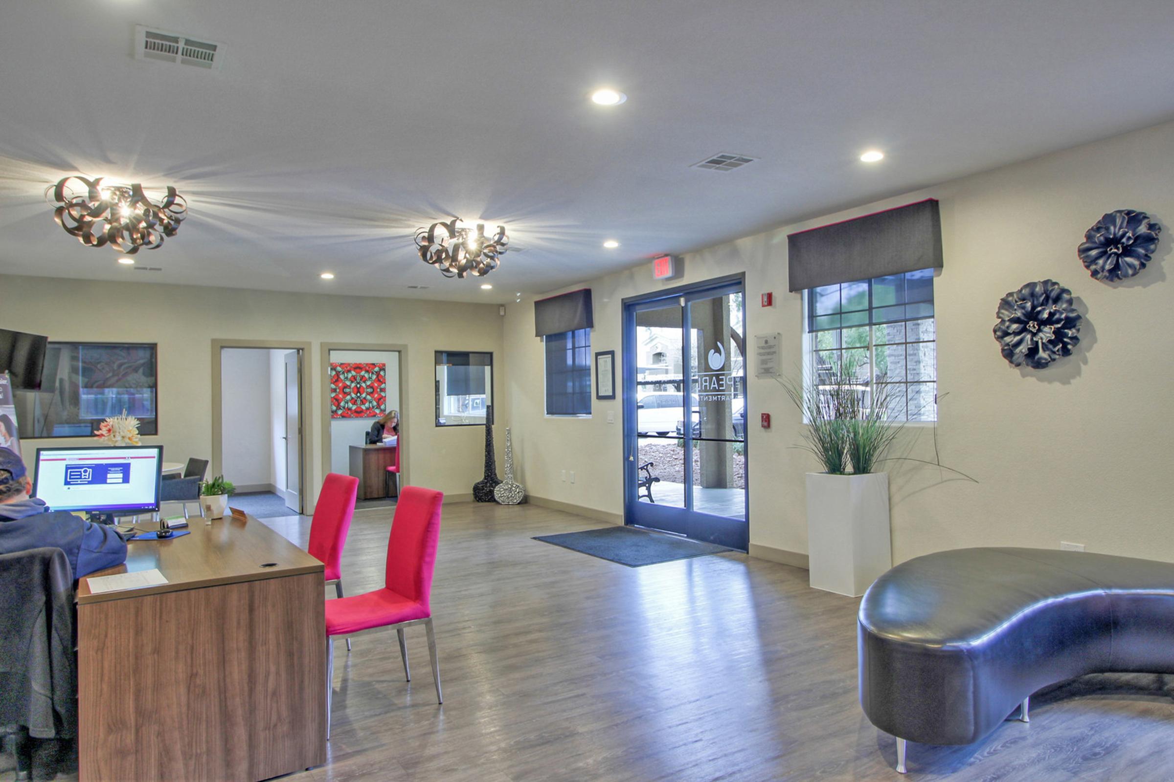 Interior of a modern reception area featuring a wooden desk with a computer, red chairs, decorative wall art, and contemporary lighting fixtures. A large glass door leads to the outside, and there are potted plants and a curved seating area, creating an inviting atmosphere.