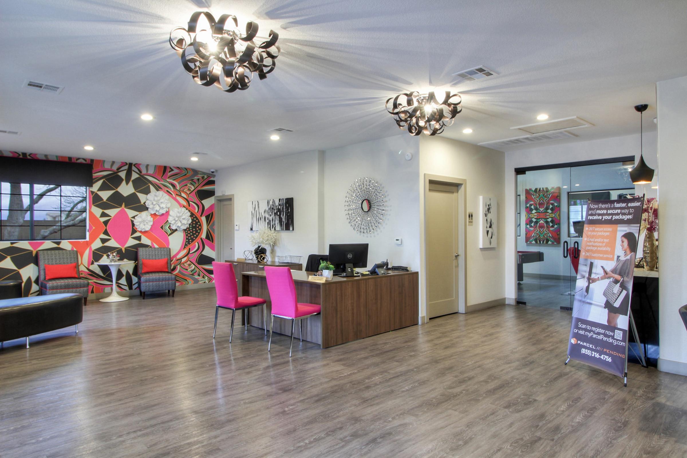 A modern reception area featuring a stylish front desk with a banner, colorful wall art, contemporary lighting fixtures, and a mix of seating options. The space has a warm and inviting atmosphere with wooden flooring and vibrant decor that adds character to the environment.