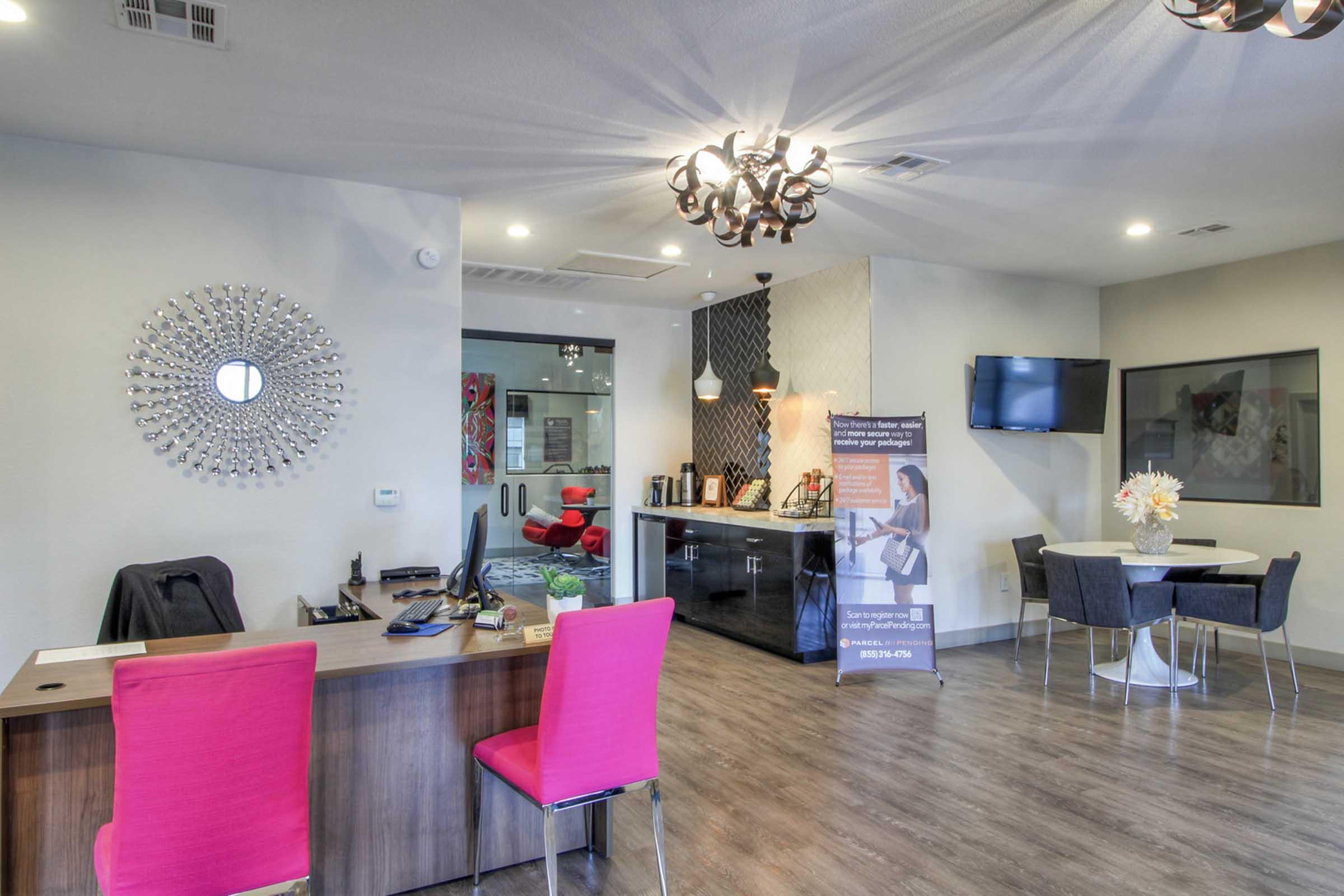 A modern reception area featuring a wooden front desk with a black phone and documents. Bright pink chairs surround a small table. A large decorative mirror and stylish lighting hang from the ceiling. A banner with text stands nearby, and a glass wall reveals a meeting space in the background. Neutral colors dominate the decor.