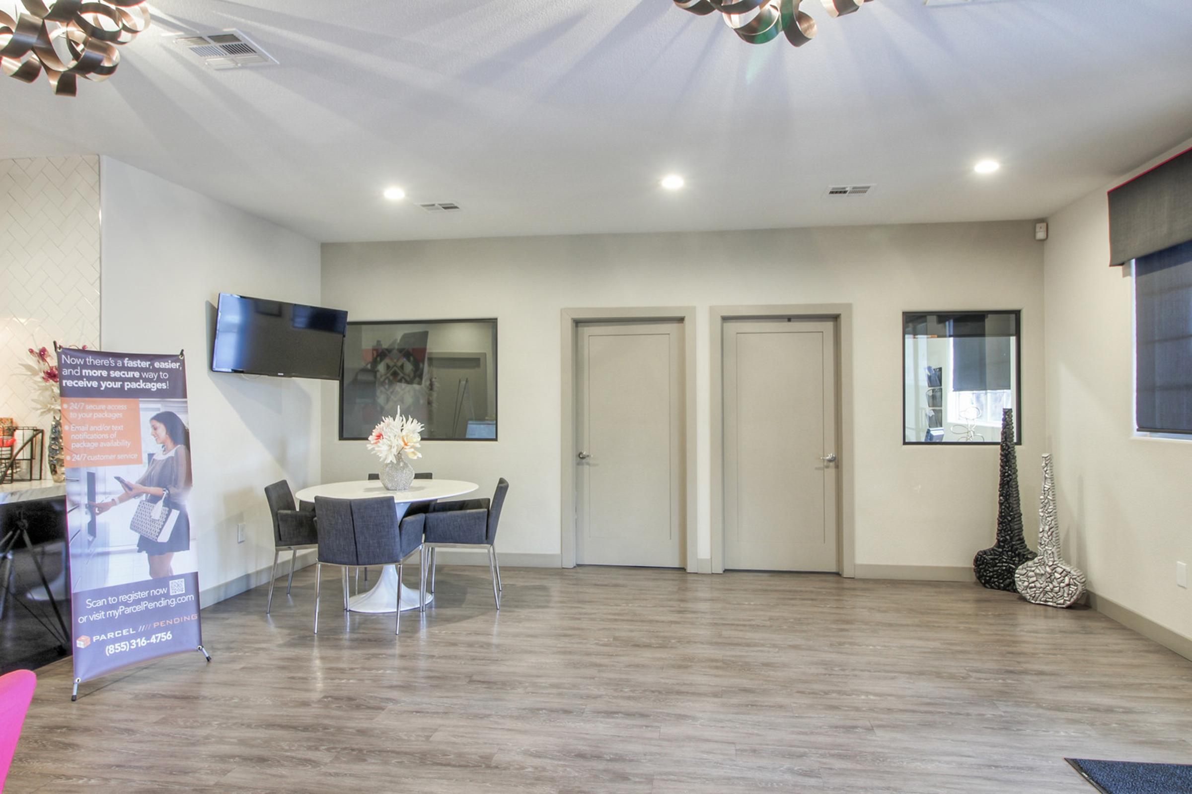 A modern interior space featuring a round dining table with chairs, a TV mounted on the wall, and two closed doors. A promotional banner is displayed in the corner, and decorative vases are present. The flooring is wooden, and the walls are painted a light color, contributing to a bright and airy atmosphere.