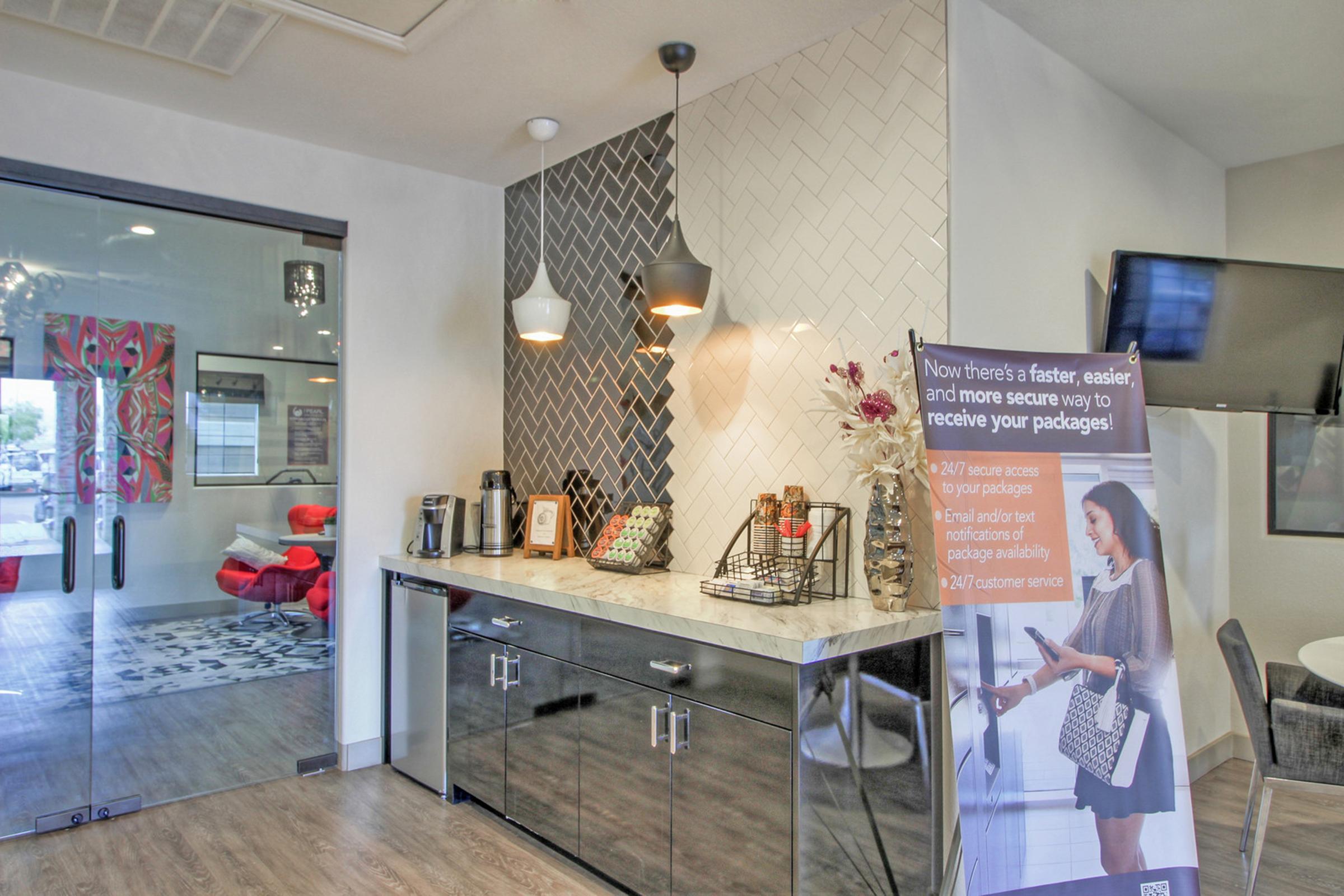 A modern reception area featuring a coffee bar with stainless steel cabinets and decorative wall tiles. There are pendant lights above the bar, and a promotional sign about package delivery services is visible. The background includes a glass-door entry leading to a brightly colored lounge area.
