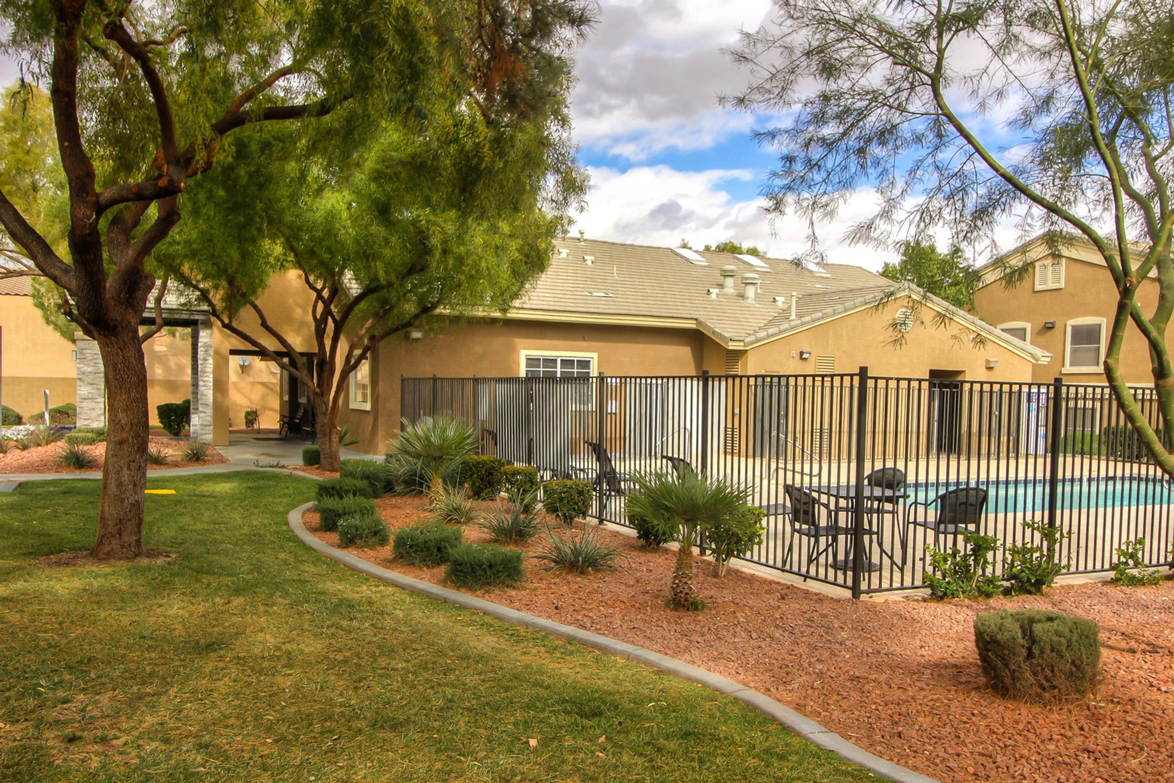 A serene outdoor scene featuring a fenced swimming pool surrounded by neatly landscaped areas. Lush green grass, decorative rocks, and various plants create a tranquil atmosphere. In the background, a multi-story building is visible under a partly cloudy sky.