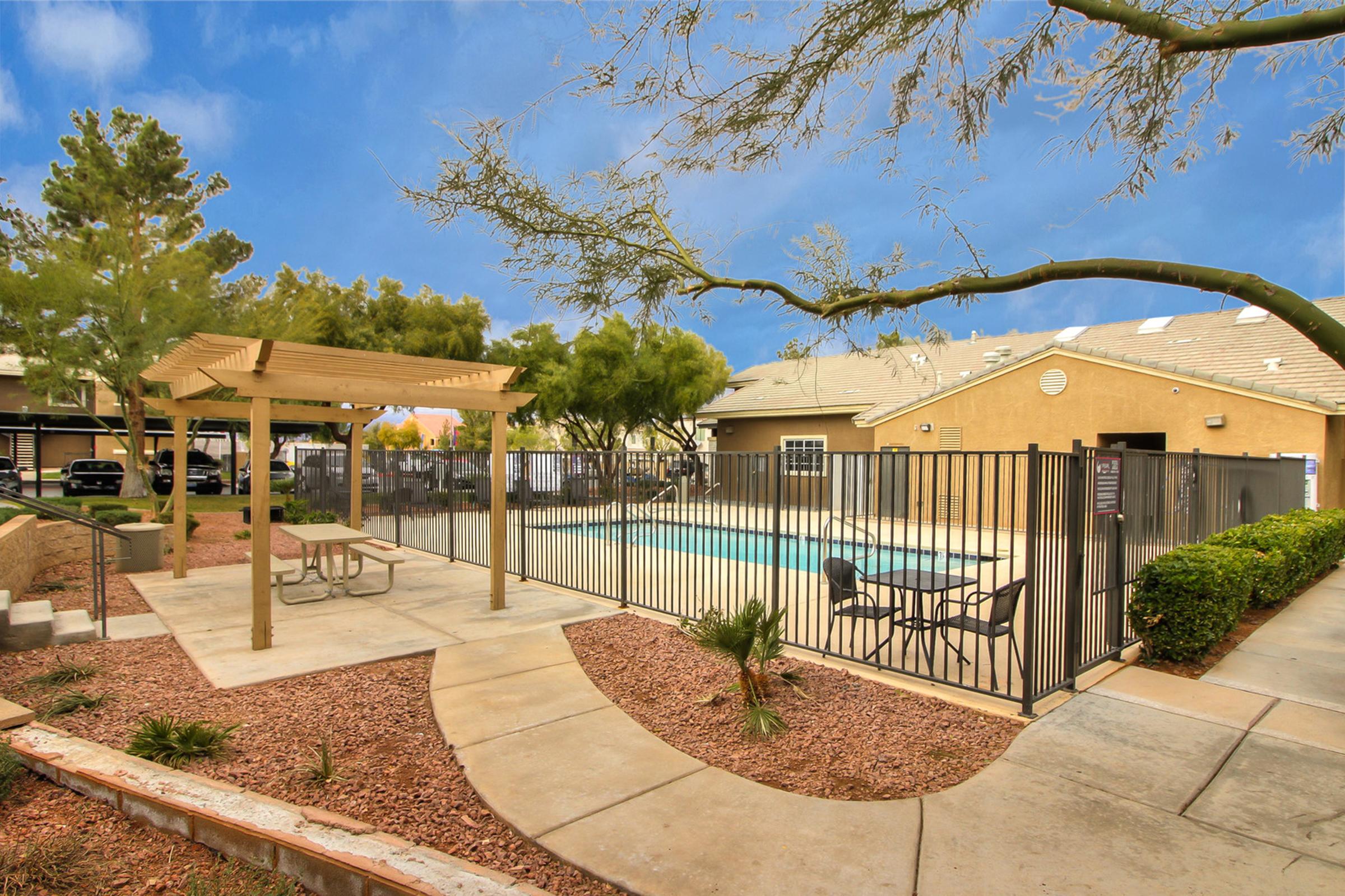A fenced swimming pool area surrounded by a landscaped outdoor space, featuring a gazebo with picnic tables, palm trees, and gravel landscaping. In the background, a residential building and a clear blue sky can be seen.