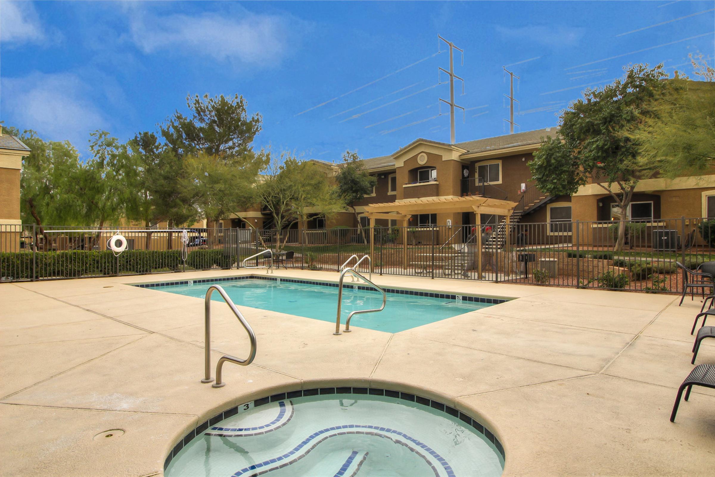 A view of a residential complex featuring a small swimming pool with a hot tub. Surrounding the pool are lounge chairs, landscaped areas with trees, and an apartment building in the background. Clear blue skies add to the inviting atmosphere of the outdoor space.