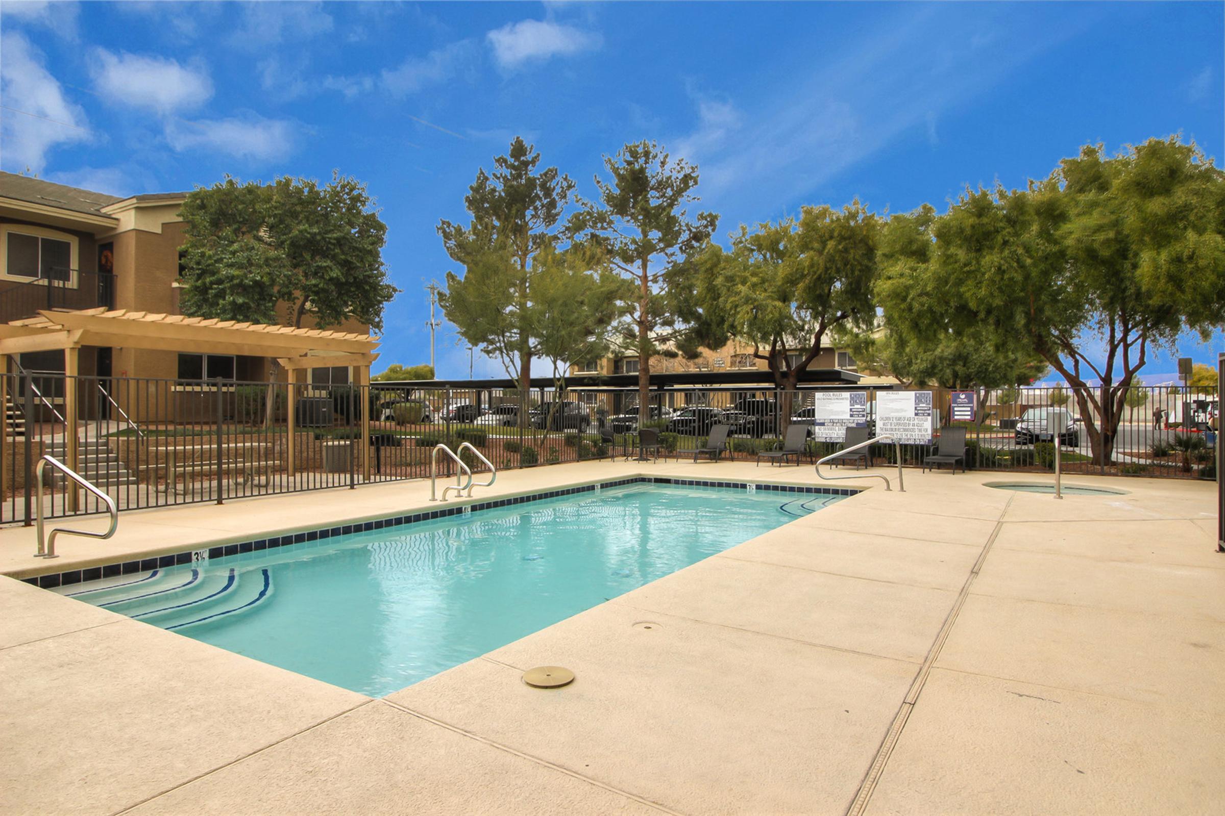 A clear, inviting swimming pool surrounded by a concrete deck and green trees. In the background, there's a pergola and seating area, creating a relaxing outdoor environment. The sky is bright blue, adding to the overall tranquil setting.
