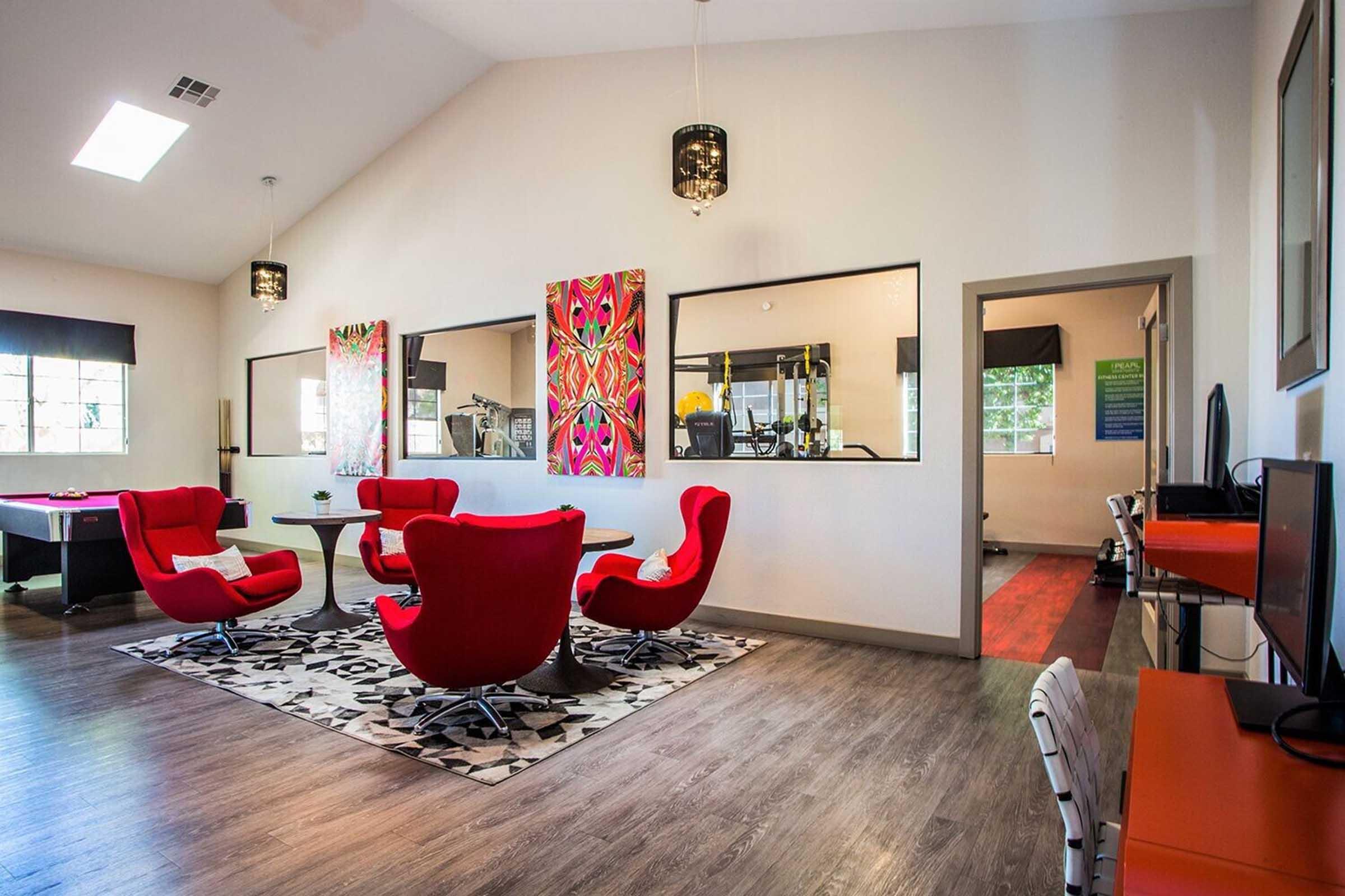 Modern communal space featuring red upholstered chairs around circular tables, a patterned area rug, and large windows allowing natural light. Two colorful abstract artworks adorn the walls. A doorway leads to a gym area, while a computer setup is visible in the background. Bright and inviting atmosphere.