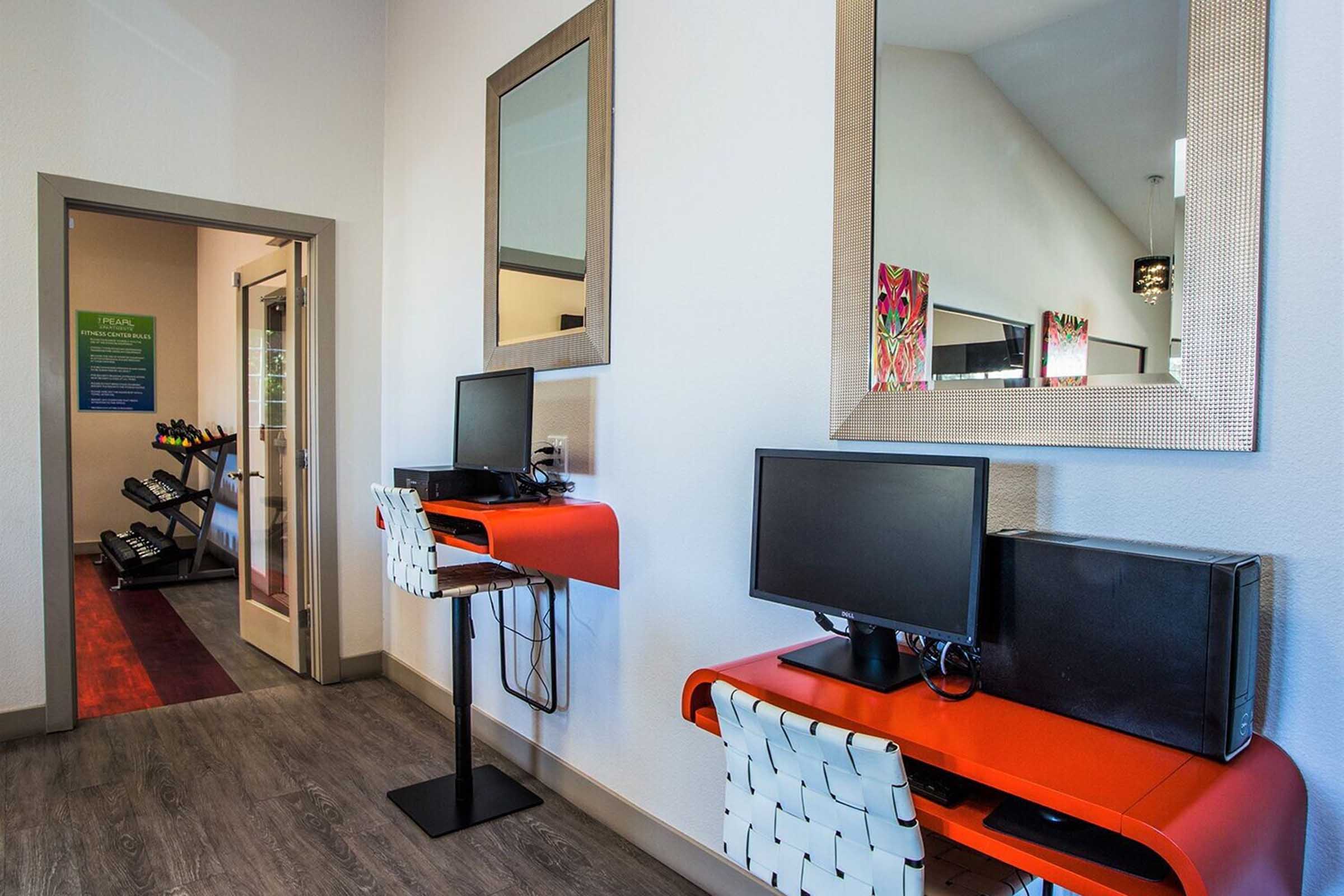 A bright, modern workspace area featuring two desktop computers on stylish red desks, with mirrors on the wall. In the background, a door leads to a fitness room equipped with exercise machines, creating a functional environment for both work and fitness.