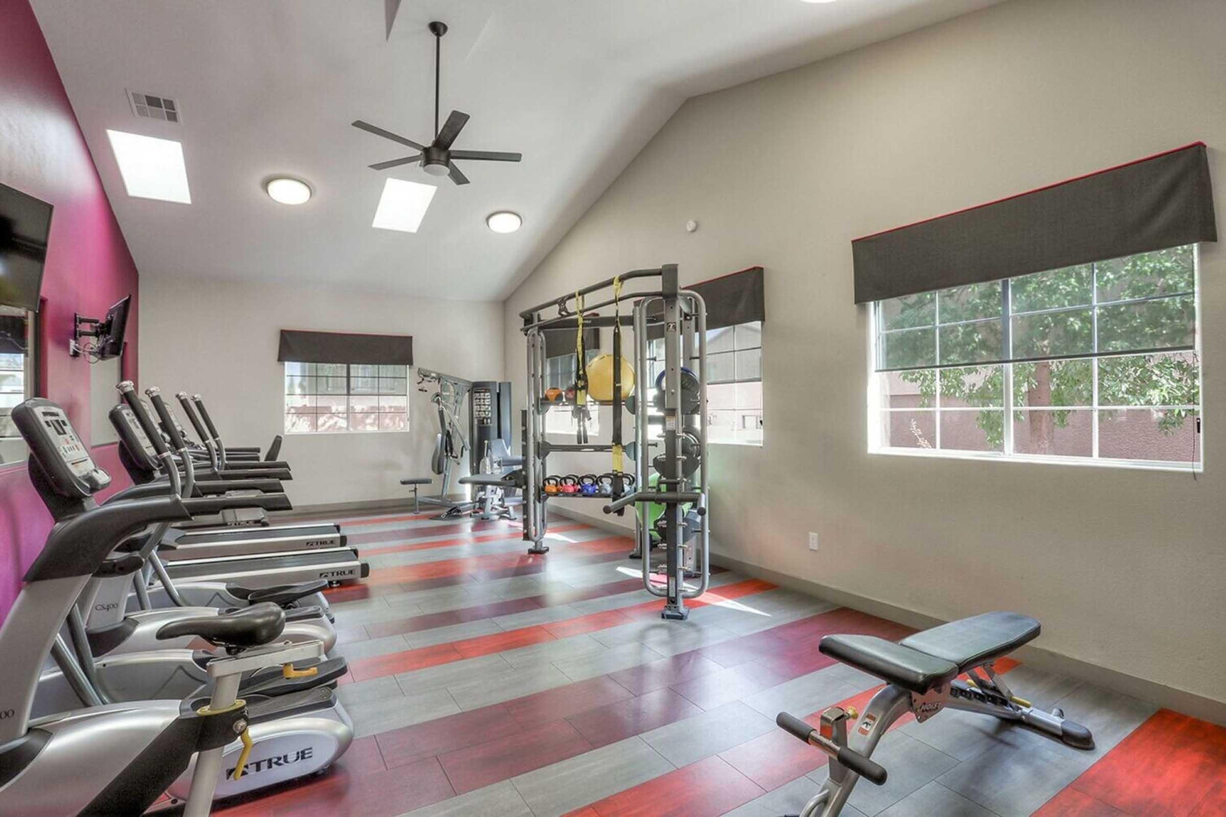 A well-lit gym with various exercise equipment, including treadmills, a weight bench, and a multifunctional strength training machine. Large windows allow natural light, and the walls are painted in neutral colors with accents. The flooring features a mix of red and black tiles for a modern look.
