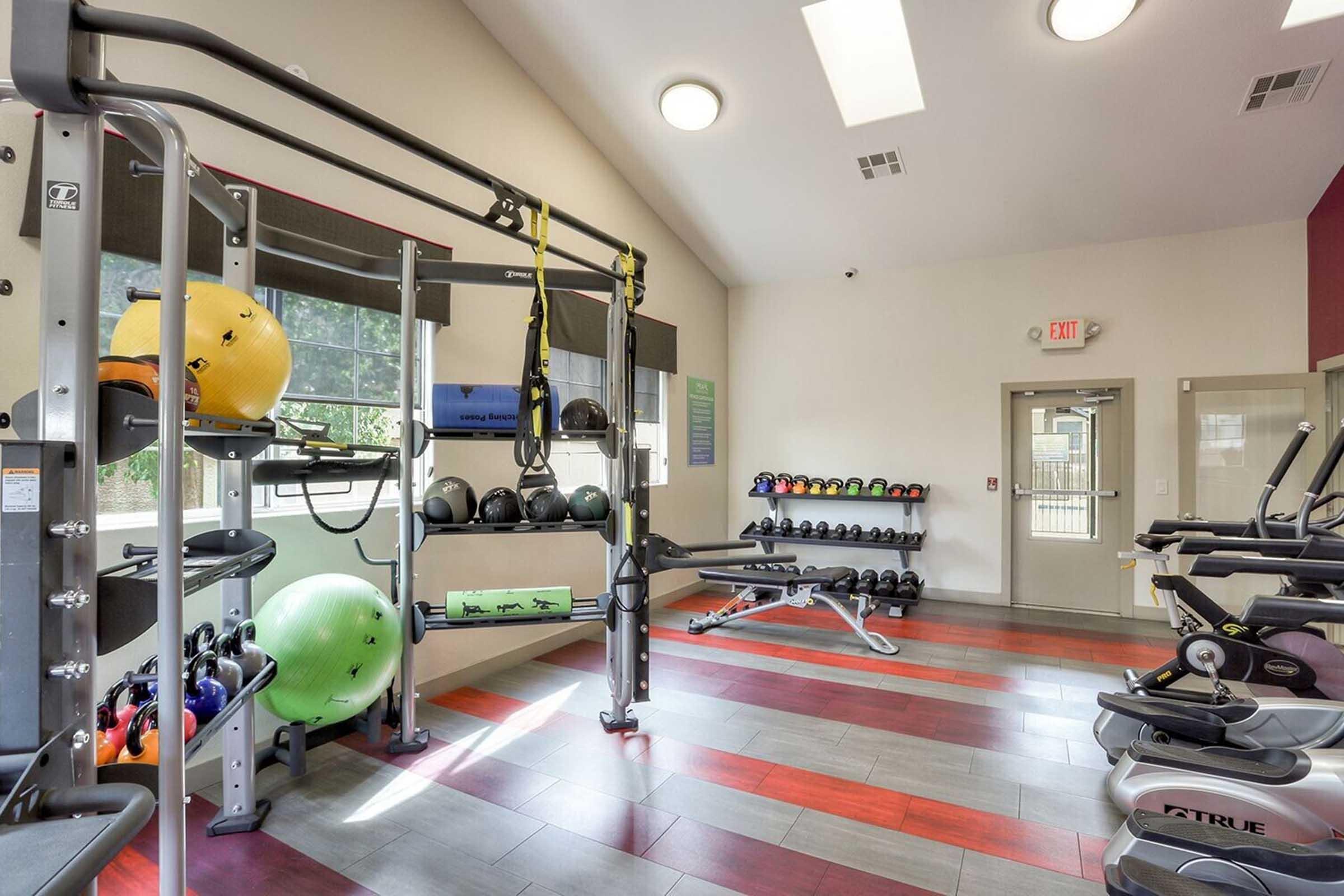 A well-lit gym with exercise equipment including weights, medicine balls, a weight bench, and cardio machines. Large windows provide natural light, and the space features modern decor with a mix of red and gray flooring.
