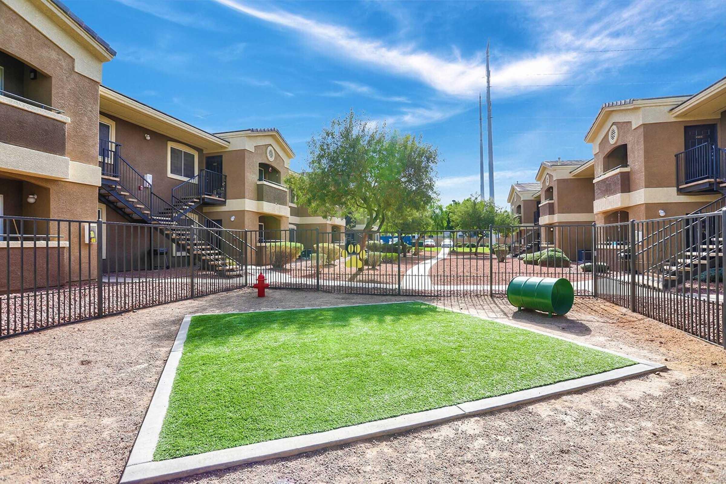 A residential apartment complex with a fenced area featuring artificial grass, a fire hydrant, and a green trash can. In the background, there are multiple two-story buildings with balconies and blue skies overhead.