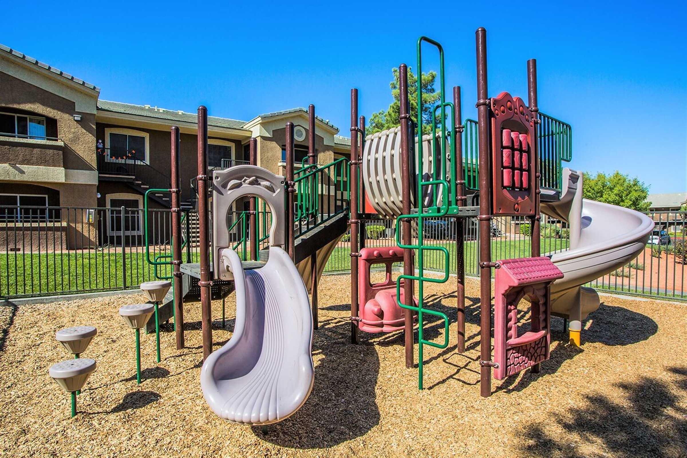 A colorful playground structure featuring slides, climbing walls, and various play features, set on a bed of wood chips. The playground is surrounded by a metal fence, with buildings visible in the background under a clear blue sky.