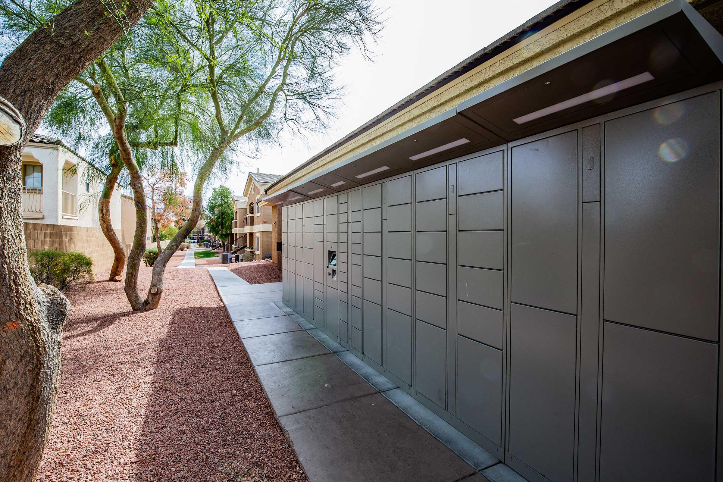 A row of grey package lockers situated along a walkway lined with gravel and trees, with residential buildings in the background. The scene is well-lit, indicating daytime, and features a clear blue sky above.