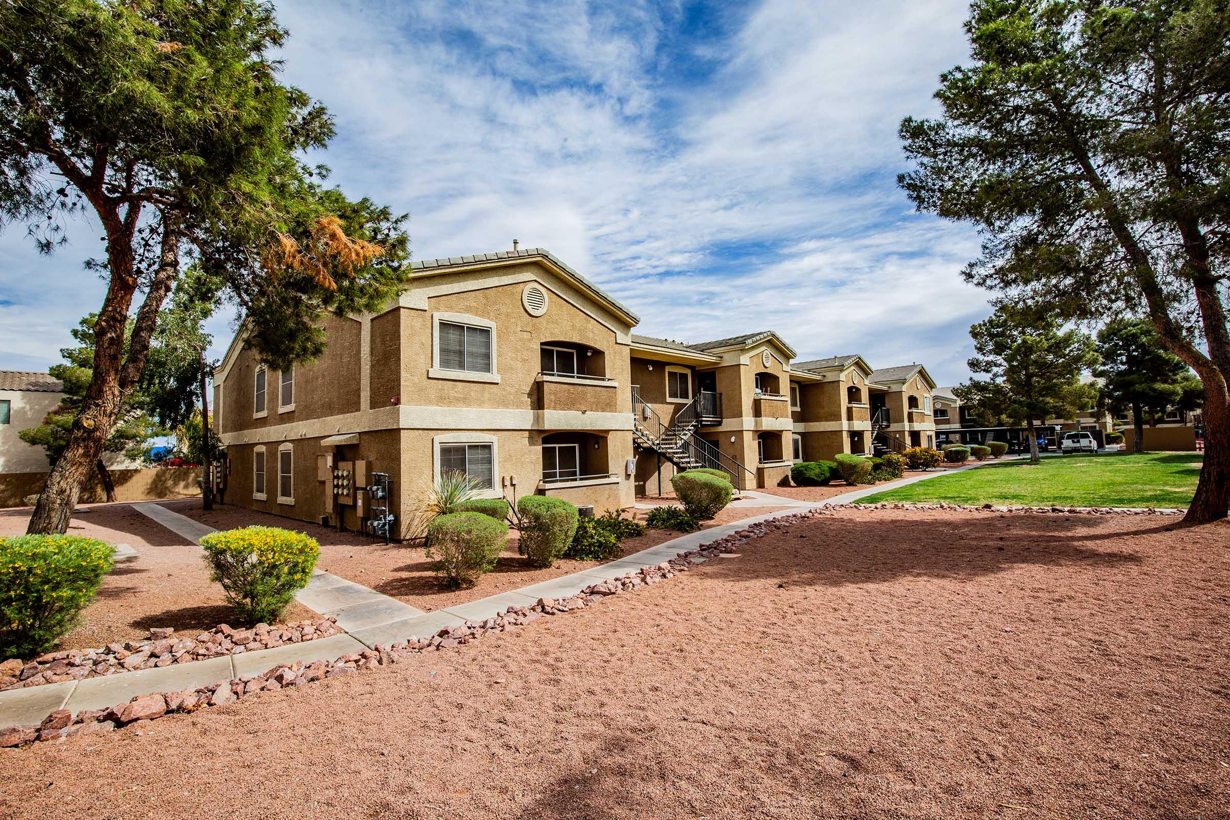 A view of an apartment complex with two-story buildings surrounded by greenery. The pathway is lined with small rocks and bushes, and there are tall trees in the foreground. The sky is partly cloudy, creating a bright and inviting atmosphere.