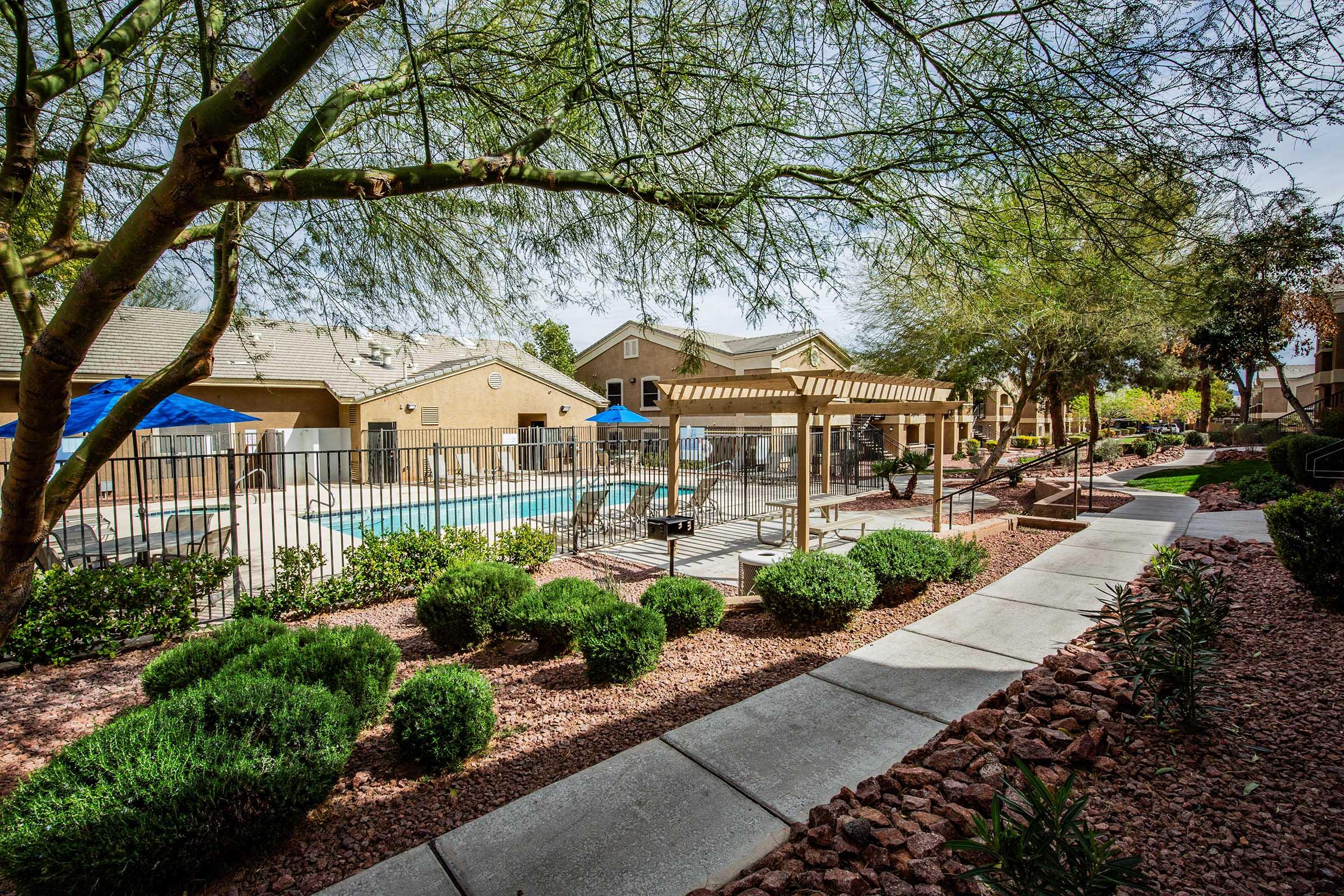 A landscaped pathway leads to a community pool area surrounded by shrubs and gravel. The pool is enclosed by a fence, with lounge chairs and umbrellas nearby. In the background, residential buildings are visible beneath a clear sky, showcasing a peaceful outdoor environment.