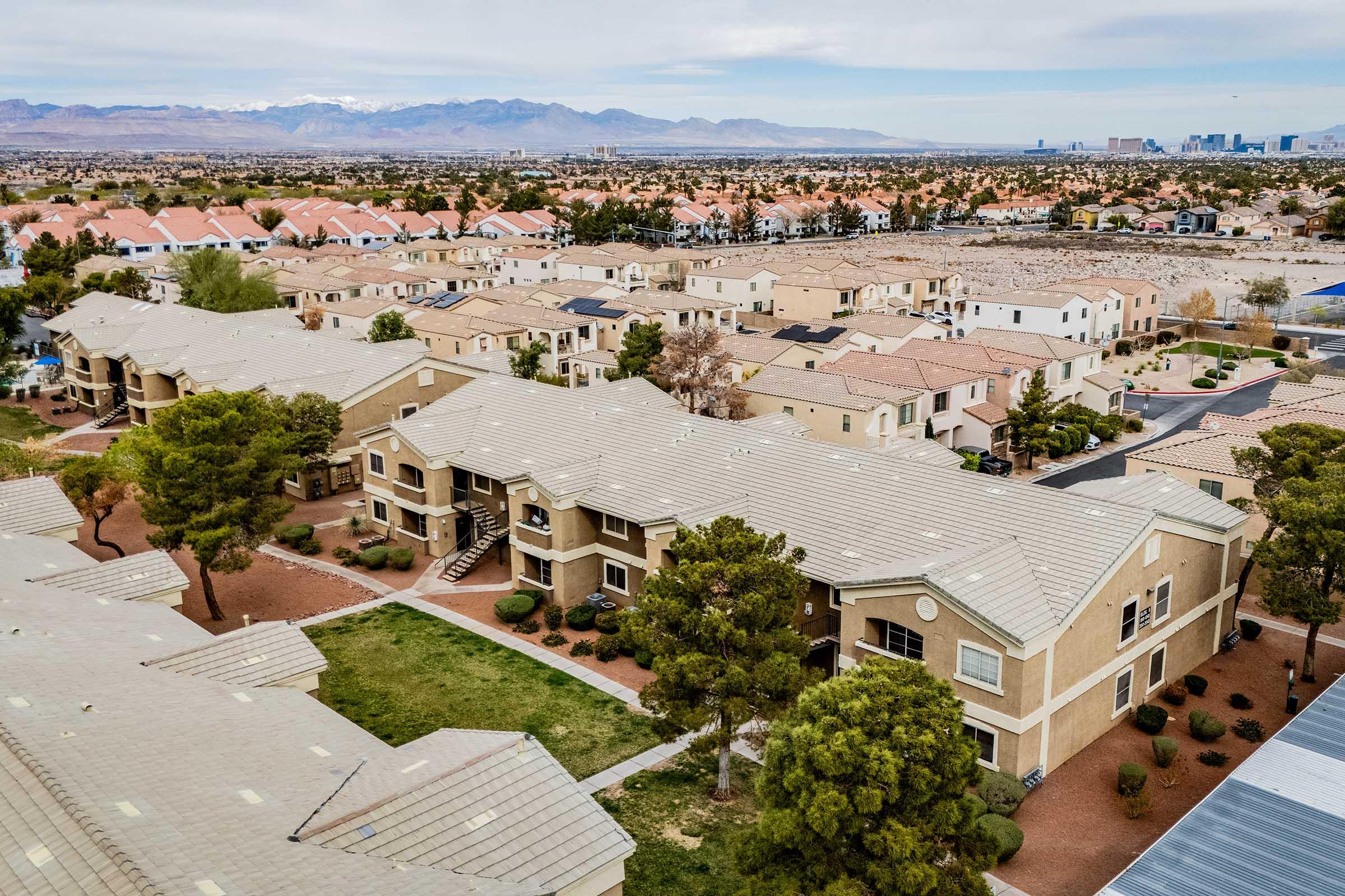 Aerial view of a residential neighborhood with beige apartment buildings and landscaped areas. In the background, low mountains and a city skyline can be seen, indicating an urban setting. The sky is partly cloudy, and the landscape features a mix of greenery and rooftops.