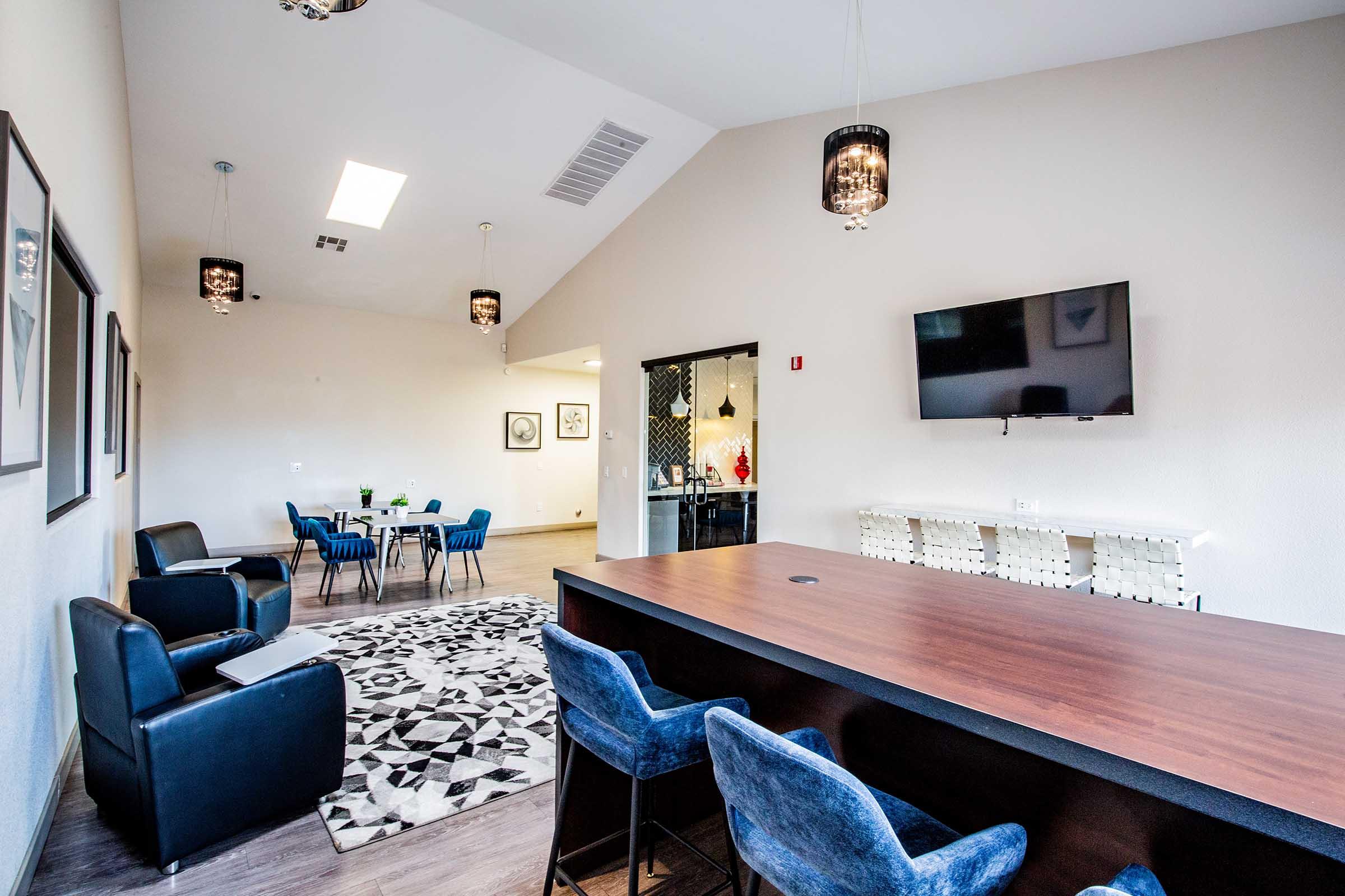 A modern interior space featuring a lounge area with blue chairs and a dark wood bar, complemented by pendant lighting. A dining table with chairs is visible in the background, and a flat-screen TV is mounted on the wall. The floor is covered in a patterned rug, and the walls are light-colored.