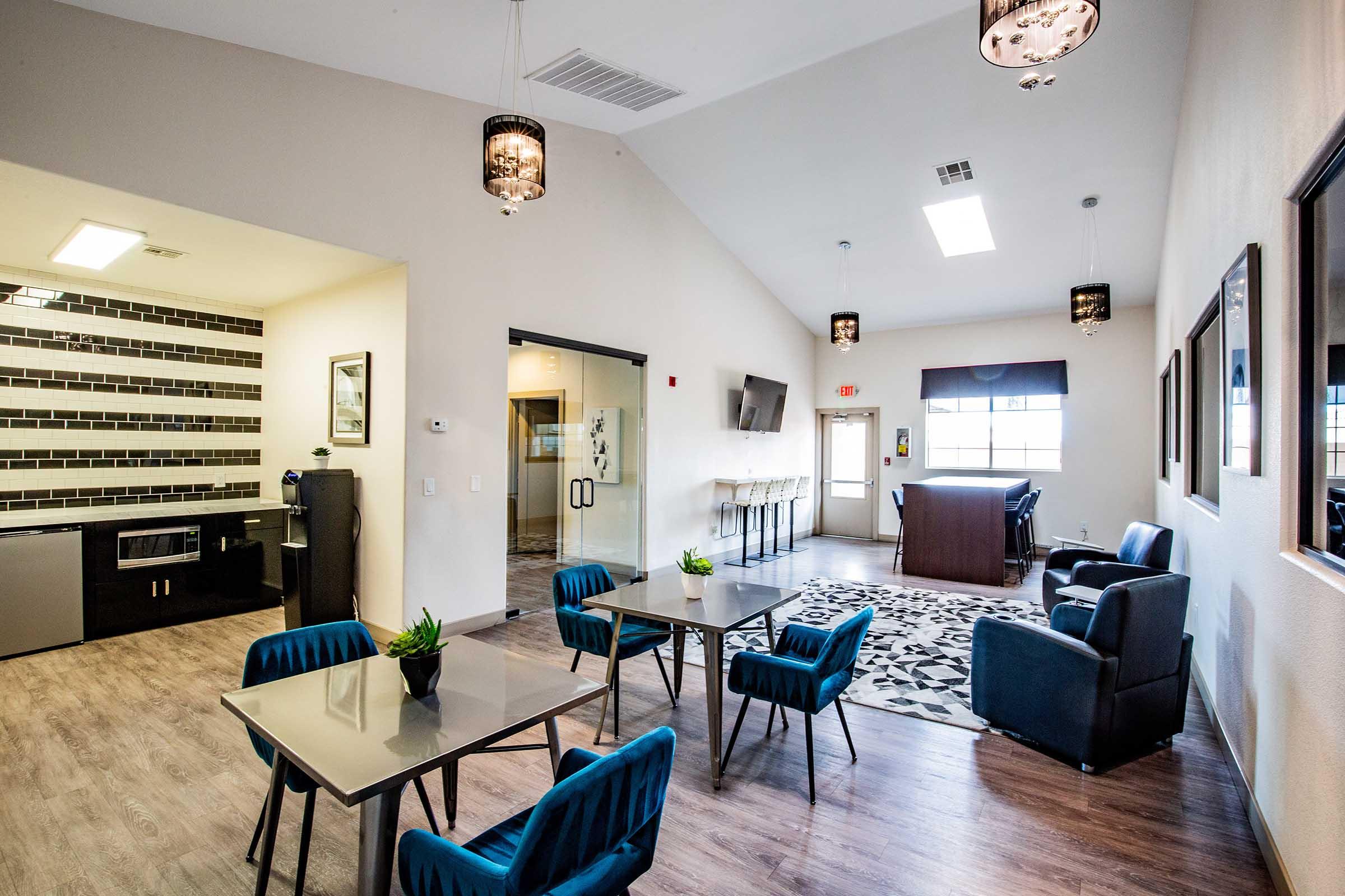 Interior view of a modern community lounge featuring stylish blue chairs and tables, a kitchenette with dark cabinets, and a large window for natural light. The space is designed with contemporary decor, including pendant lighting and a patterned area rug. A bar-height table is visible in the background.