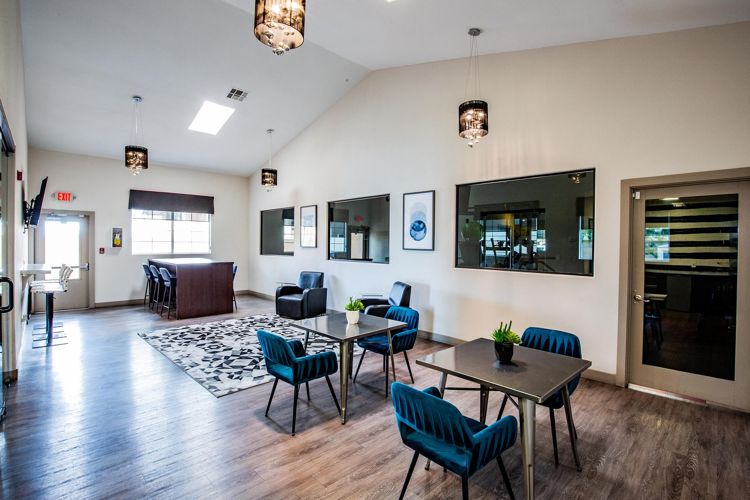 A spacious interior of a modern lounge area featuring dark wood furniture, a bar counter, and several seating arrangements with blue chairs and tables. The room has high ceilings, large windows, and decorative light fixtures, creating a bright and inviting atmosphere.
