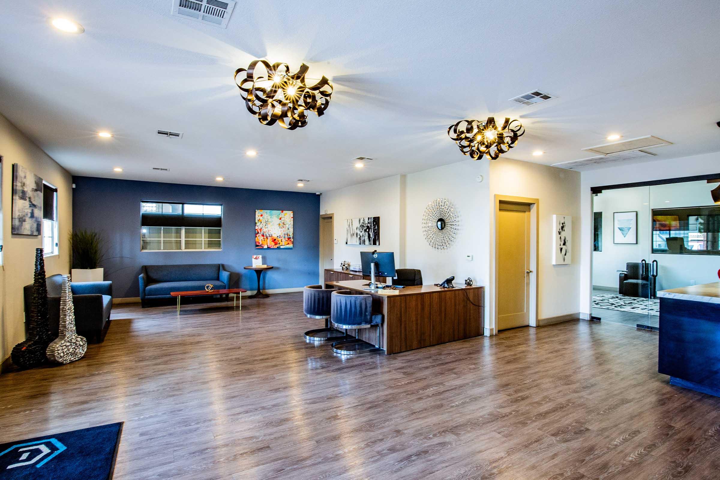 A modern office interior featuring stylish light fixtures, sleek wooden flooring, and contemporary furniture. The space includes a reception area with a wooden desk, seating for visitors, and decorative wall art. A separate lounge area is visible in the background.