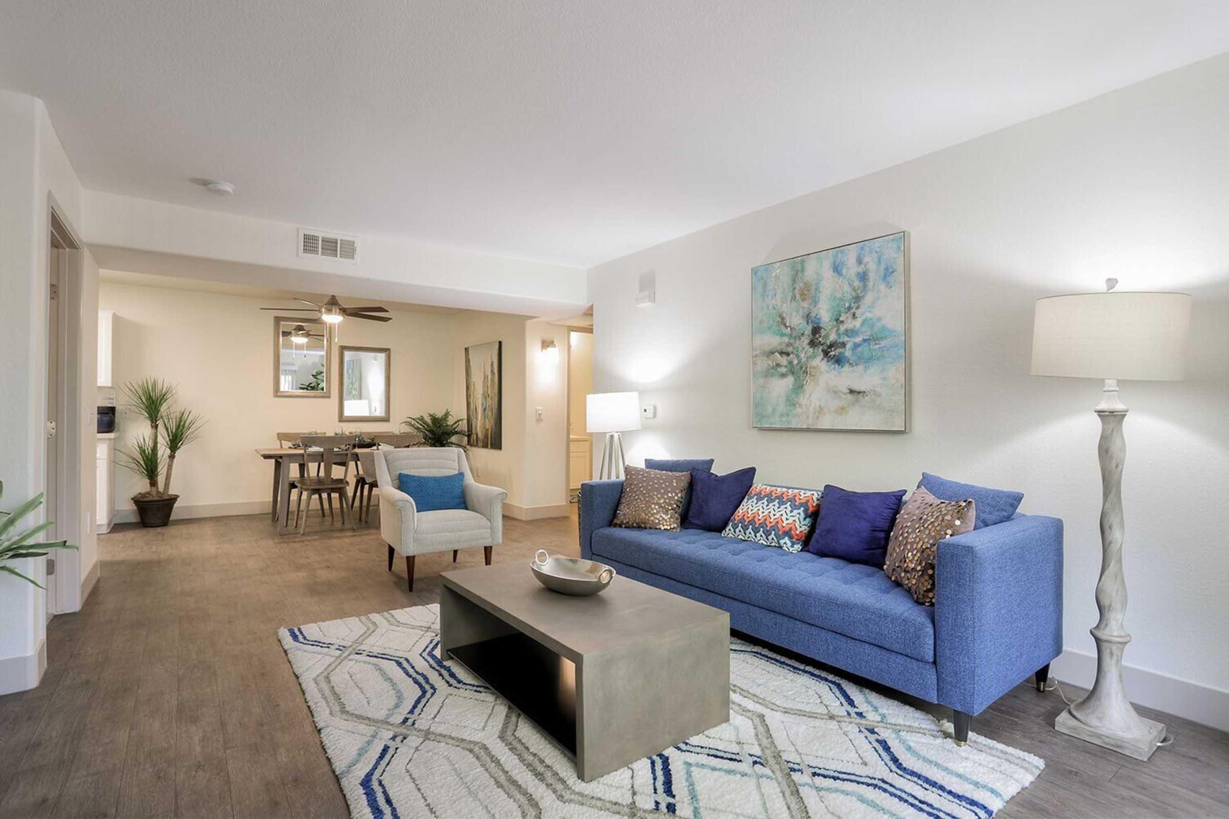 A modern living room featuring a blue sofa with colorful cushions, a coffee table, and a light-colored rug with geometric patterns. There are potted plants, a floor lamp, and artwork on the walls. In the background, there's a dining area with a wooden table and chairs, illuminated by a ceiling fan.