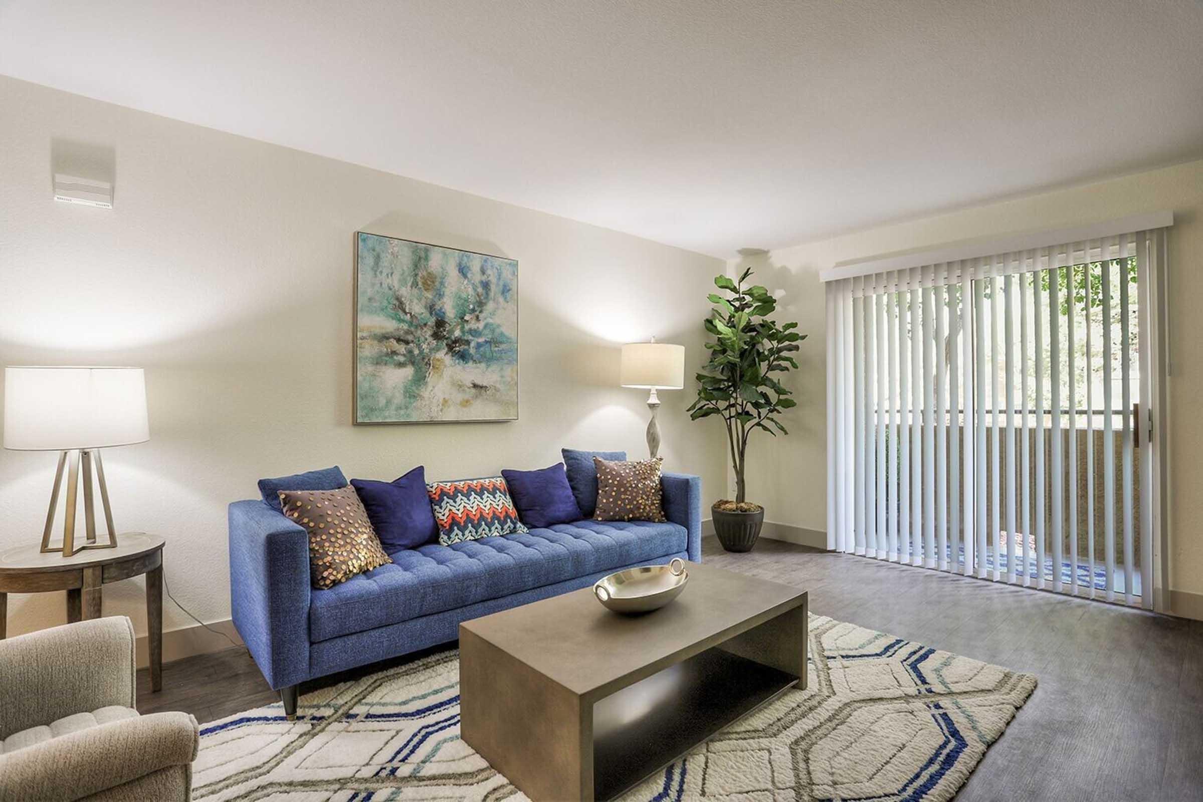 A cozy living room featuring a blue sofa with decorative pillows, a coffee table, and a stylish rug. A standing lamp and a small side table are present, along with a large potted plant. The room has sliding glass doors with vertical blinds, allowing natural light to enter and offering a view of the outdoors.