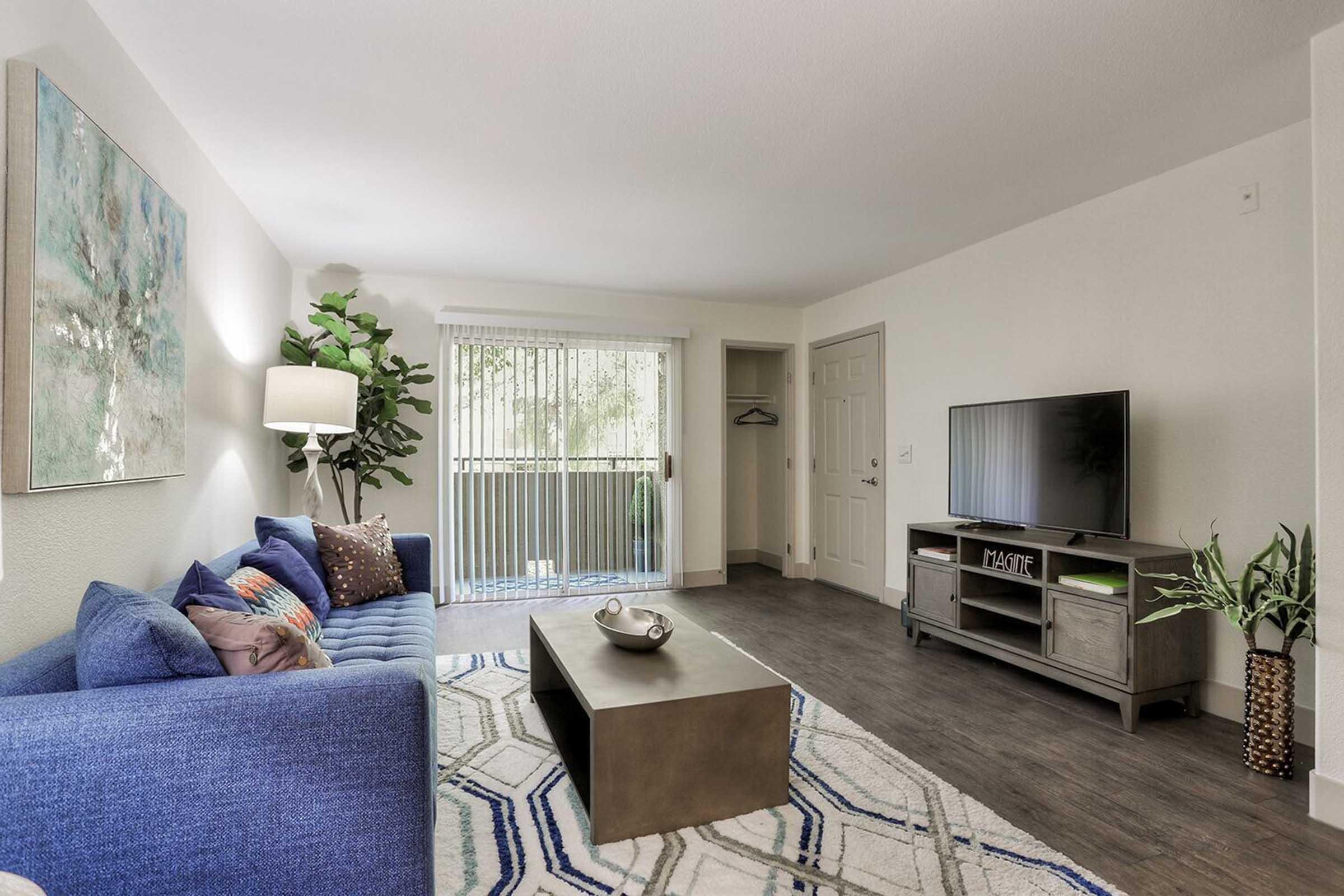 A bright, modern living room featuring a blue sofa with decorative pillows, a coffee table, and a television on a stand. Natural light fills the space, highlighting a nearby potted plant and a piece of wall art. A sliding door leads to a small balcony. The flooring is dark wood, contrasting with the light walls.