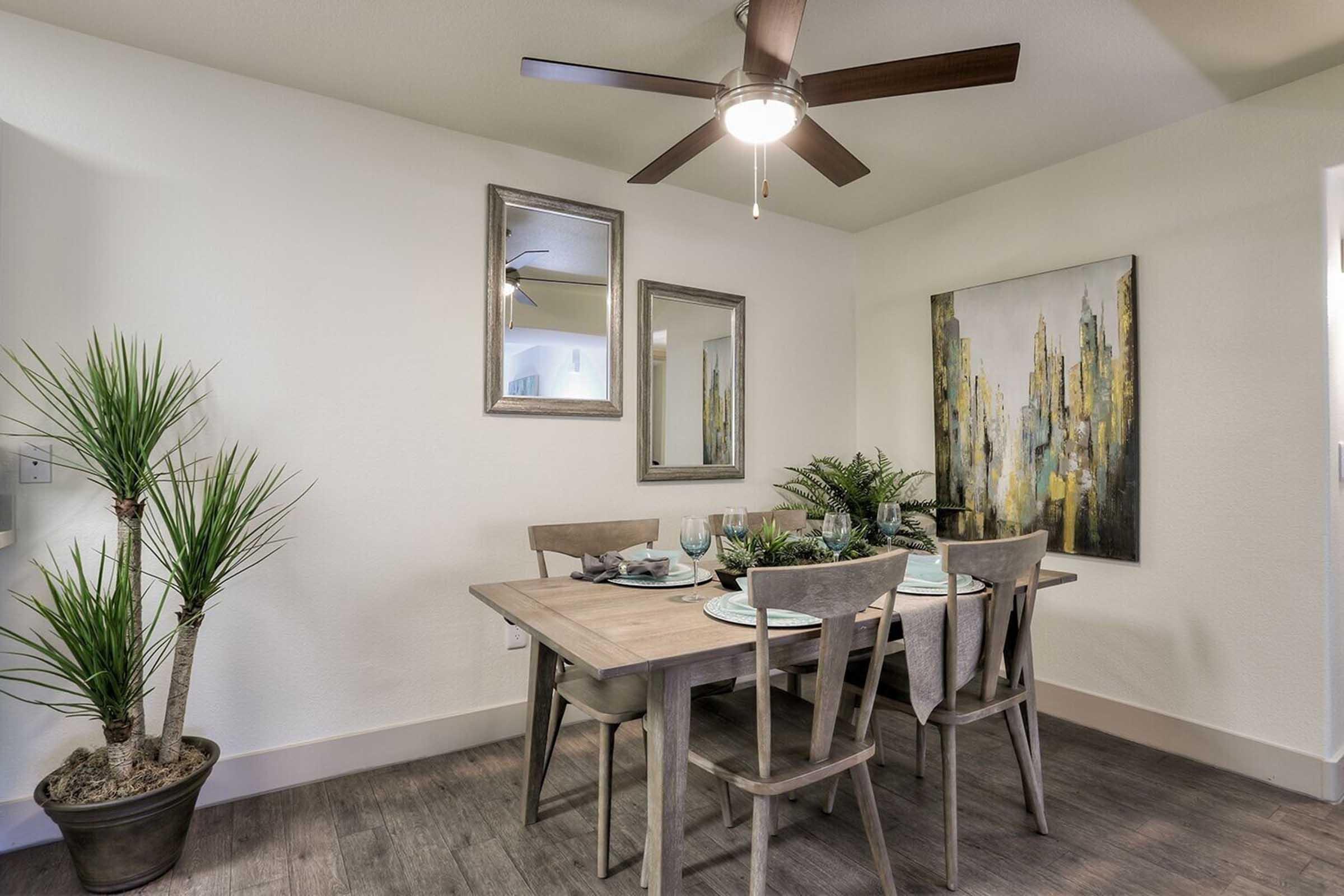A modern dining area featuring a light wooden table set for four, with stylish tableware. There are two framed mirrors on the wall, a large abstract painting with yellow and gray tones, and a potted plant in the corner, all in a well-lit space with a ceiling fan.