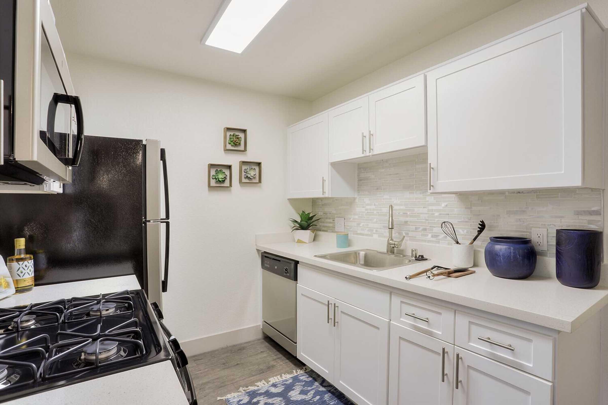 Modern kitchen featuring white cabinets, a stainless steel sink, and appliances including a microwave, dishwasher, and gas stove. The countertop is light-colored with a decorative blue item and plants on the wall. There are small framed pictures on the wall above the sink. Bright overhead lighting enhances the space.
