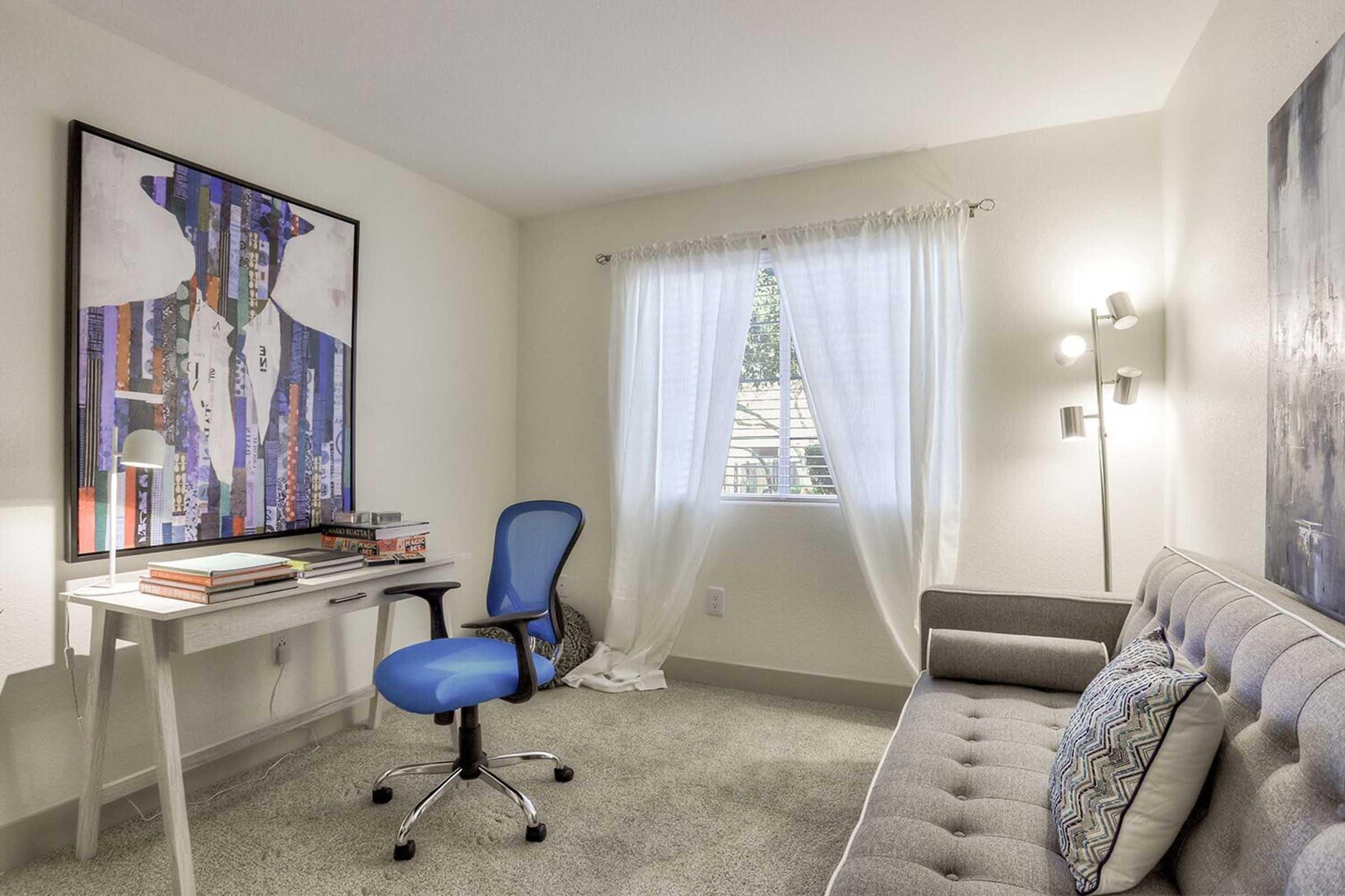 A cozy room featuring a gray sofa, a desk with a lamp, and a blue ergonomic chair. A colorful abstract painting hangs on the wall, while a large window with sheer white curtains allows natural light to illuminate the space. The carpeted floor adds warmth to the modern aesthetic.