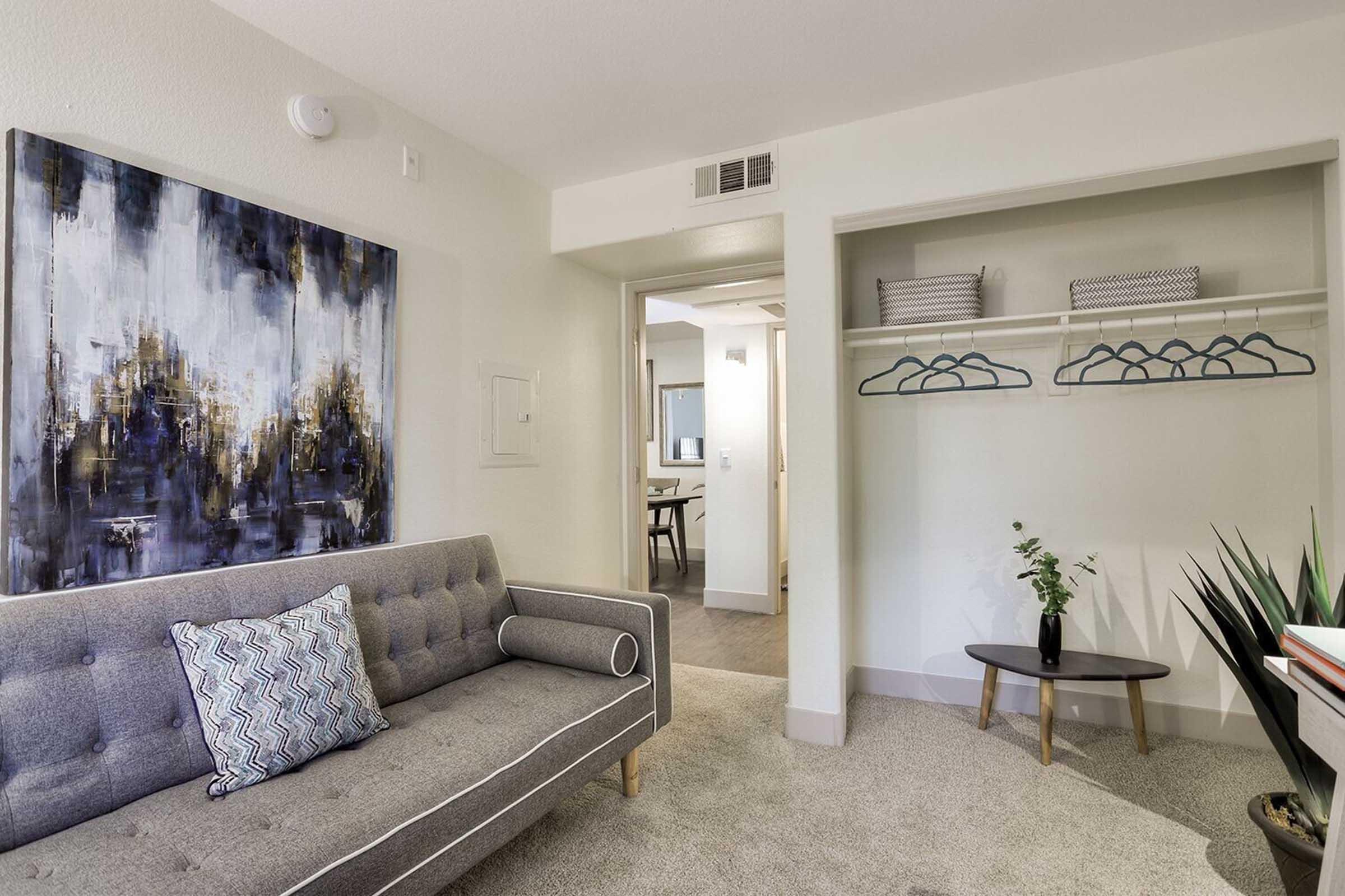 A modern living space featuring a tufted gray sofa with a decorative pillow, a colorful abstract painting on the wall, and an open closet with hanging shelves and blue hangers. A small round table and a potted plant add character to the room. The design is bright and inviting.