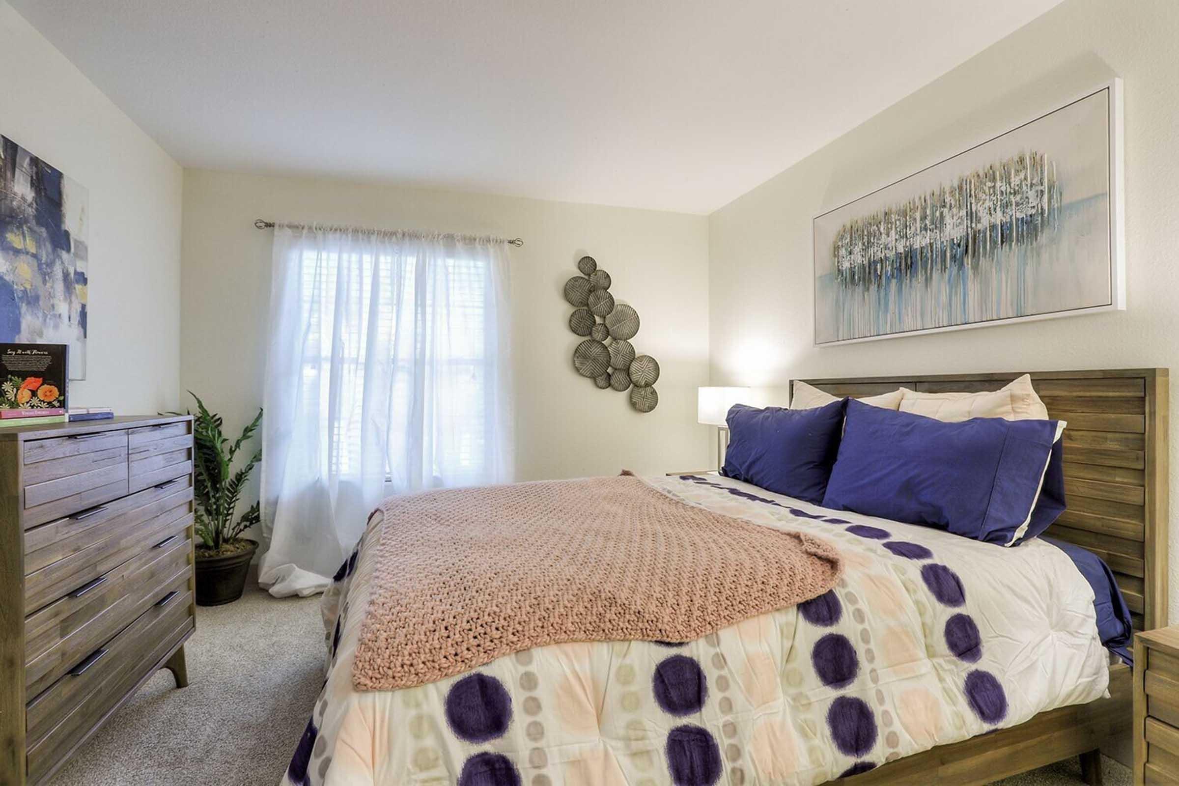 A cozy bedroom featuring a wooden bed with purple and white bedding, a chunky pink throw blanket, and decorative pillows. There's a dresser with a piece of artwork above it, a large window with sheer curtains allowing natural light, a potted plant, and a modern painting on the wall.