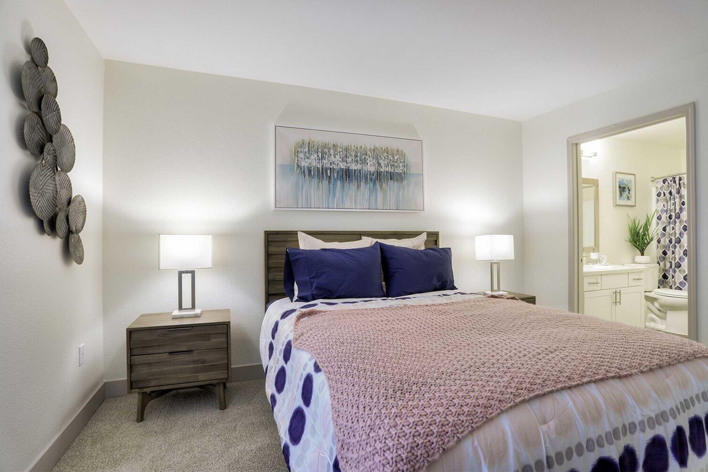 Cozy bedroom featuring a queen-sized bed with a pink and white patterned throw blanket, two bedside lamps, and a decorative wall art piece. A light-colored nightstand sits next to the bed. In the background, a bathroom is partially visible with a plant and colorful shower curtain.