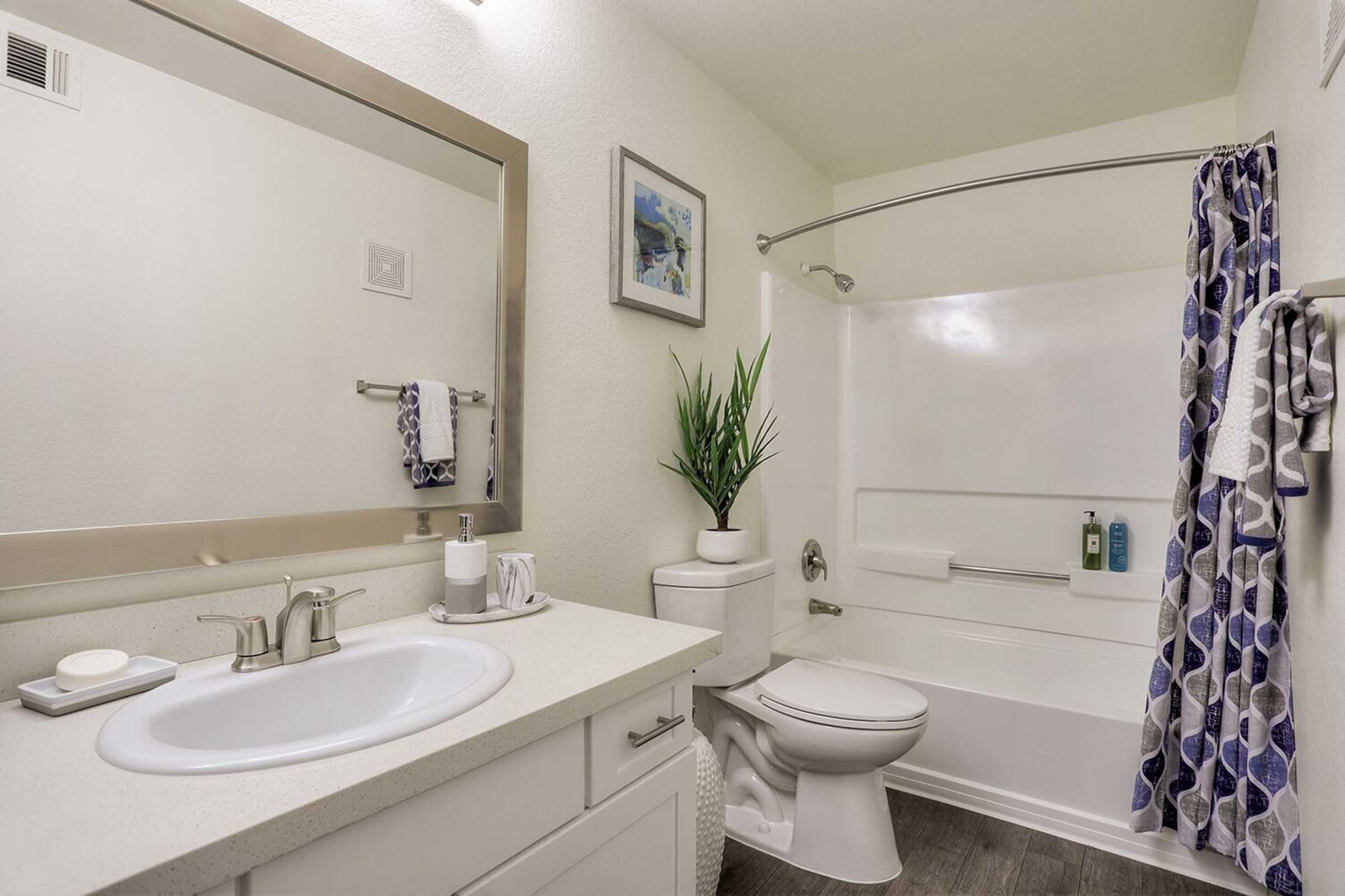 A clean and modern bathroom featuring a white sink with a mirror above, a white toilet, and a bathtub with a shower. There is a potted plant, towels hanging on a rack, and toiletries on the edge of the bathtub. The walls are painted pale, contributing to a bright and airy atmosphere.