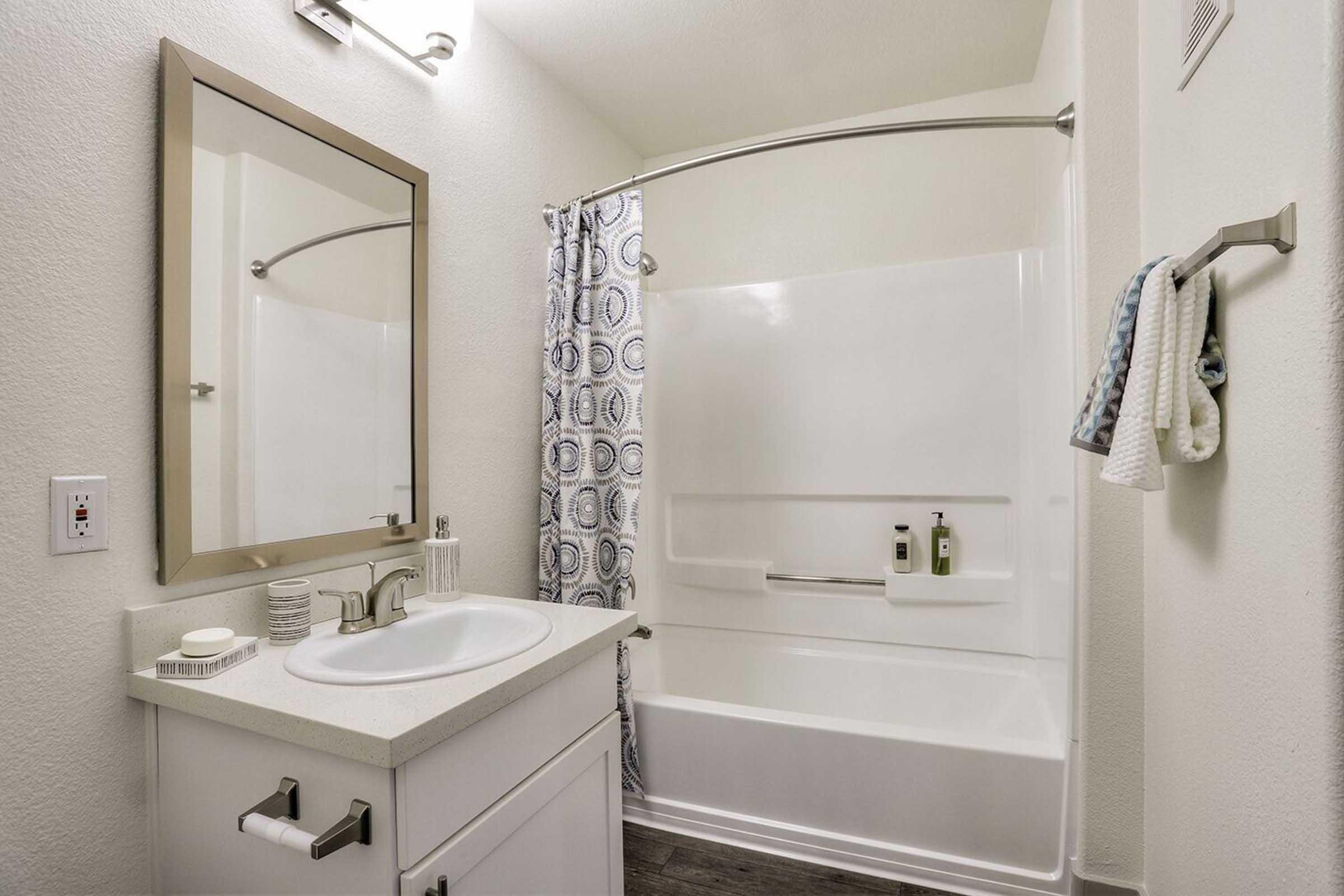 A clean, modern bathroom featuring a white bathtub with a curved shower curtain rod, a mirror above a white sink with storage drawers, and a decorative towel hanging on a bar. The walls are painted in a light color, and there are minimalist decorations and toiletries on the sink counter.