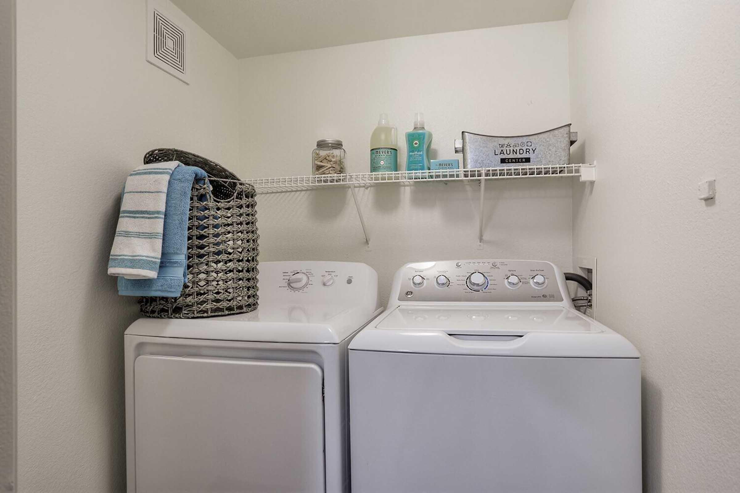 A laundry area featuring a white washing machine and dryer side by side. Above them, a shelf holds laundry supplies including bottles and a bin labeled "Laundry." A folded blue towel hangs on the side of the dryer. The walls are painted a light color, creating a clean and organized appearance.