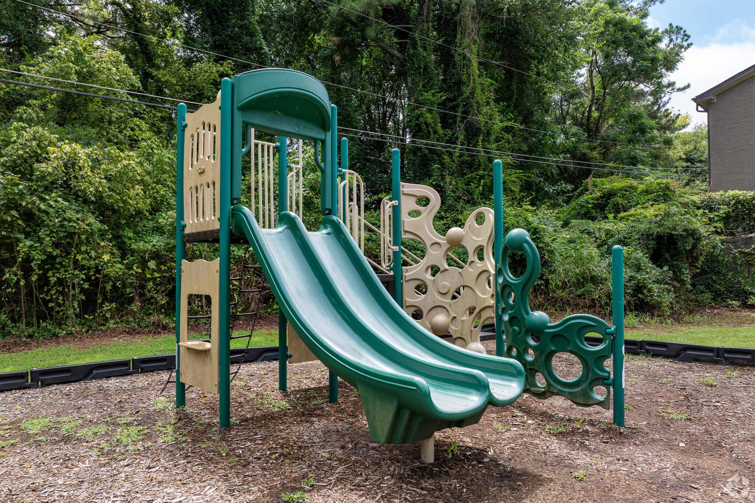 A playground featuring two green slides, climbing structures, and various play panels, surrounded by lush greenery and trees. The ground is covered with wood chips, and there are no children playing in the area. The design is modern and colorful, emphasizing outdoor play.
