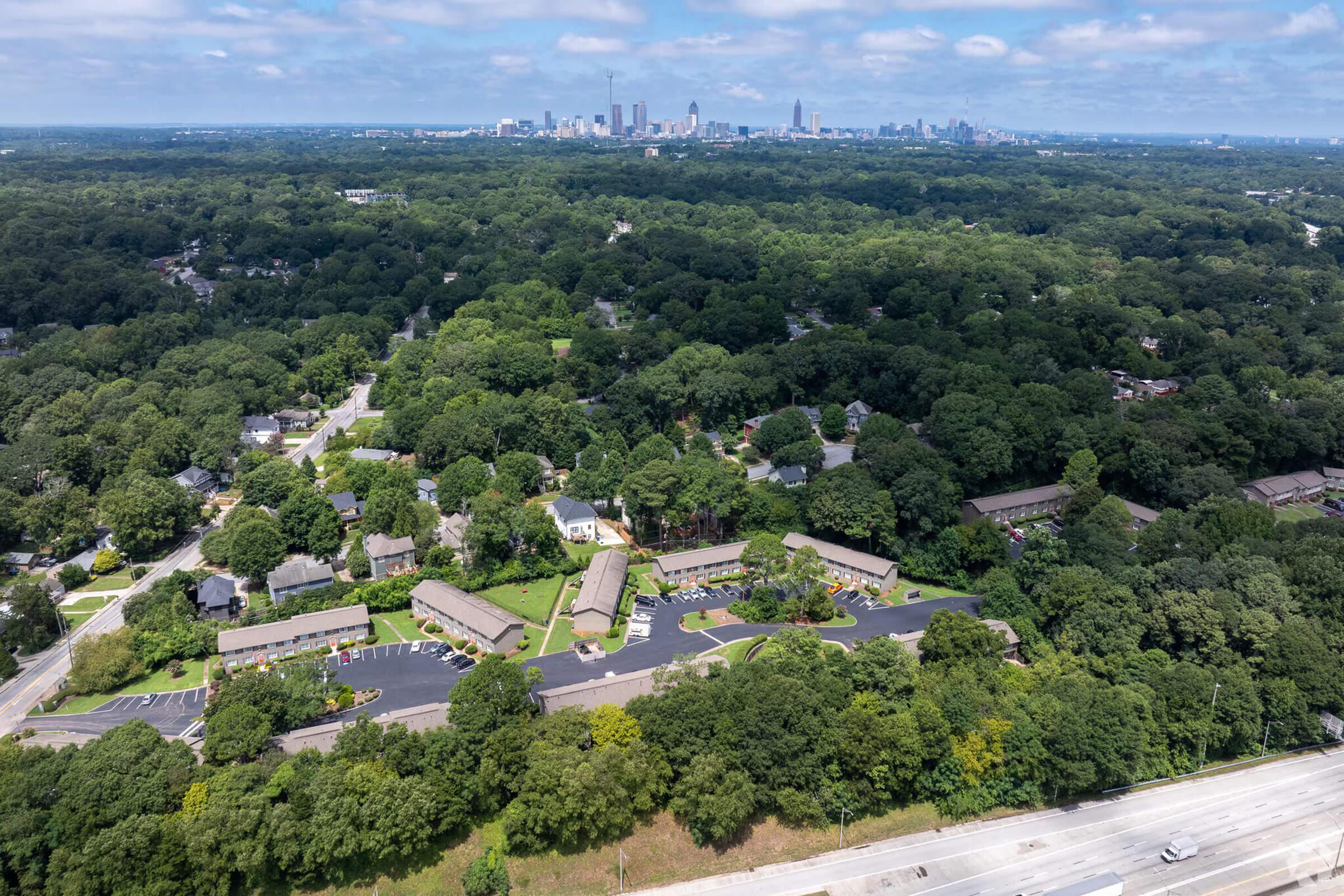 Aerial view of a suburban neighborhood surrounded by greenery, with low-rise residential buildings in the foreground. In the distance, a city skyline is visible under a partly cloudy sky. The scene contrasts natural landscapes with urban architecture.