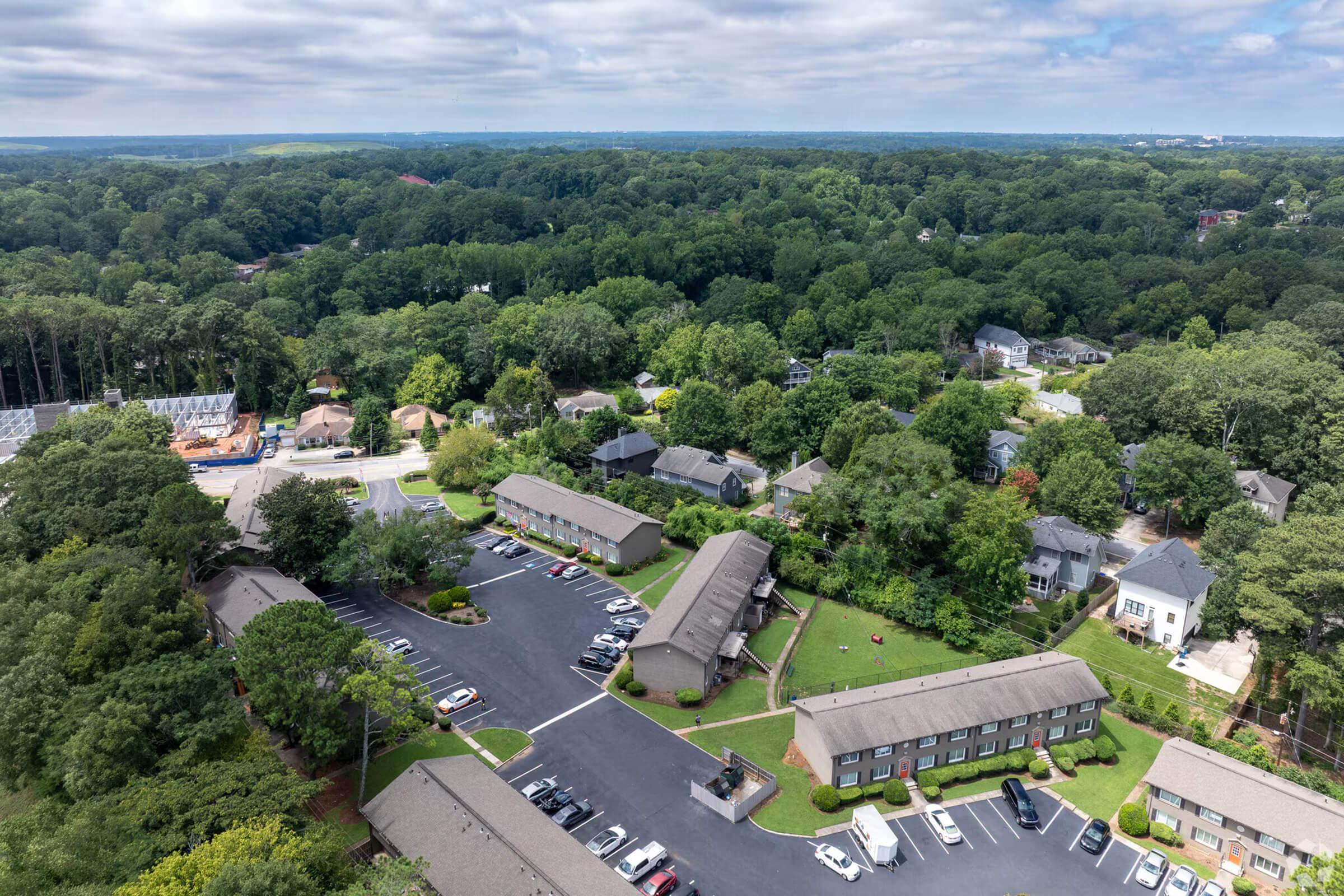 Aerial view of a suburban area featuring multiple residential buildings, parking lots, and green trees. The scene shows a mix of townhouses and apartments, surrounded by lush foliage and roads, under a partly cloudy sky.