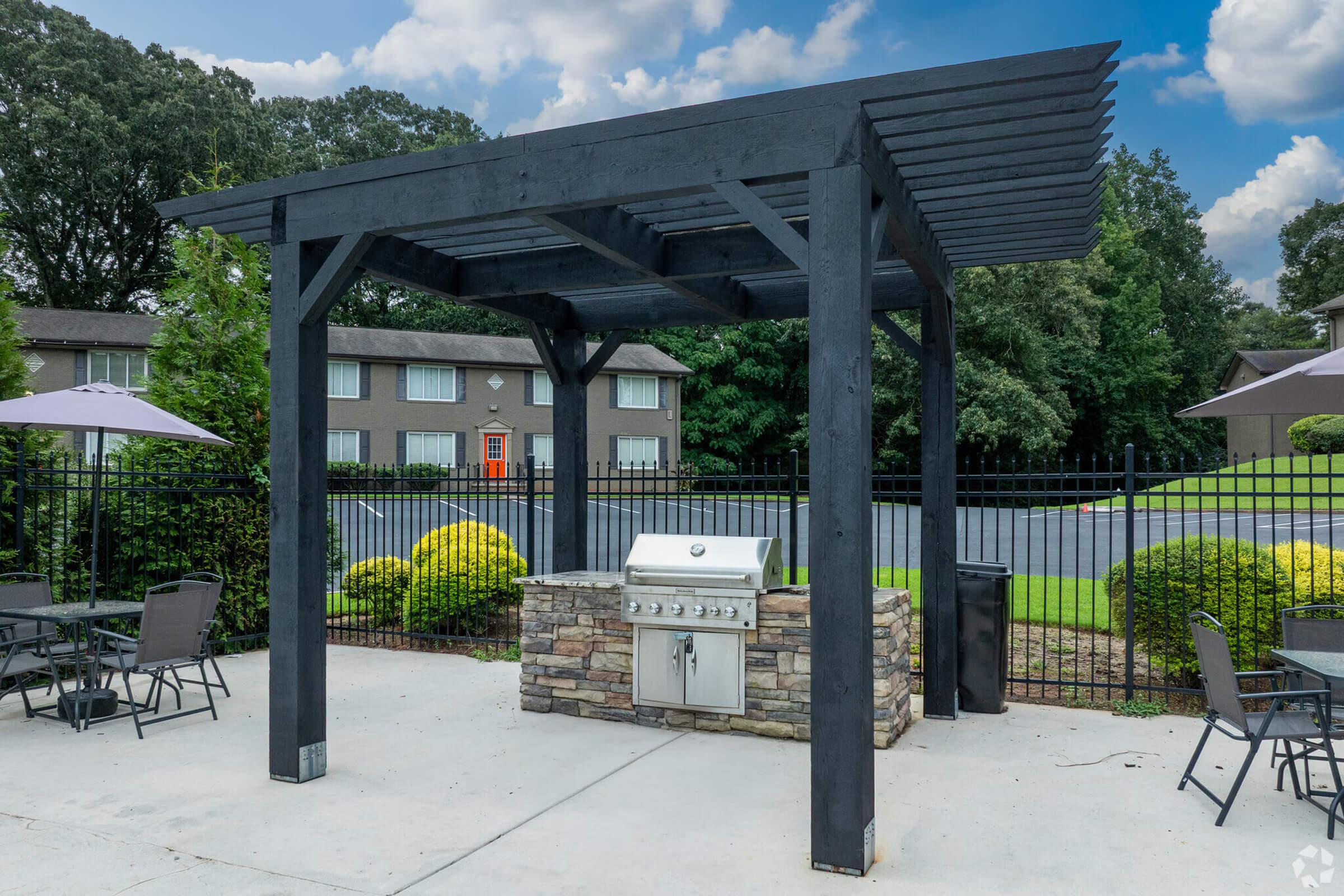 A covered outdoor grilling area with a stone barbecue grill set on a concrete patio. Surrounding the grill are metal tables and chairs under umbrellas, with well-maintained greenery and a fence in the background. Trees and an apartment building are visible in the scene.