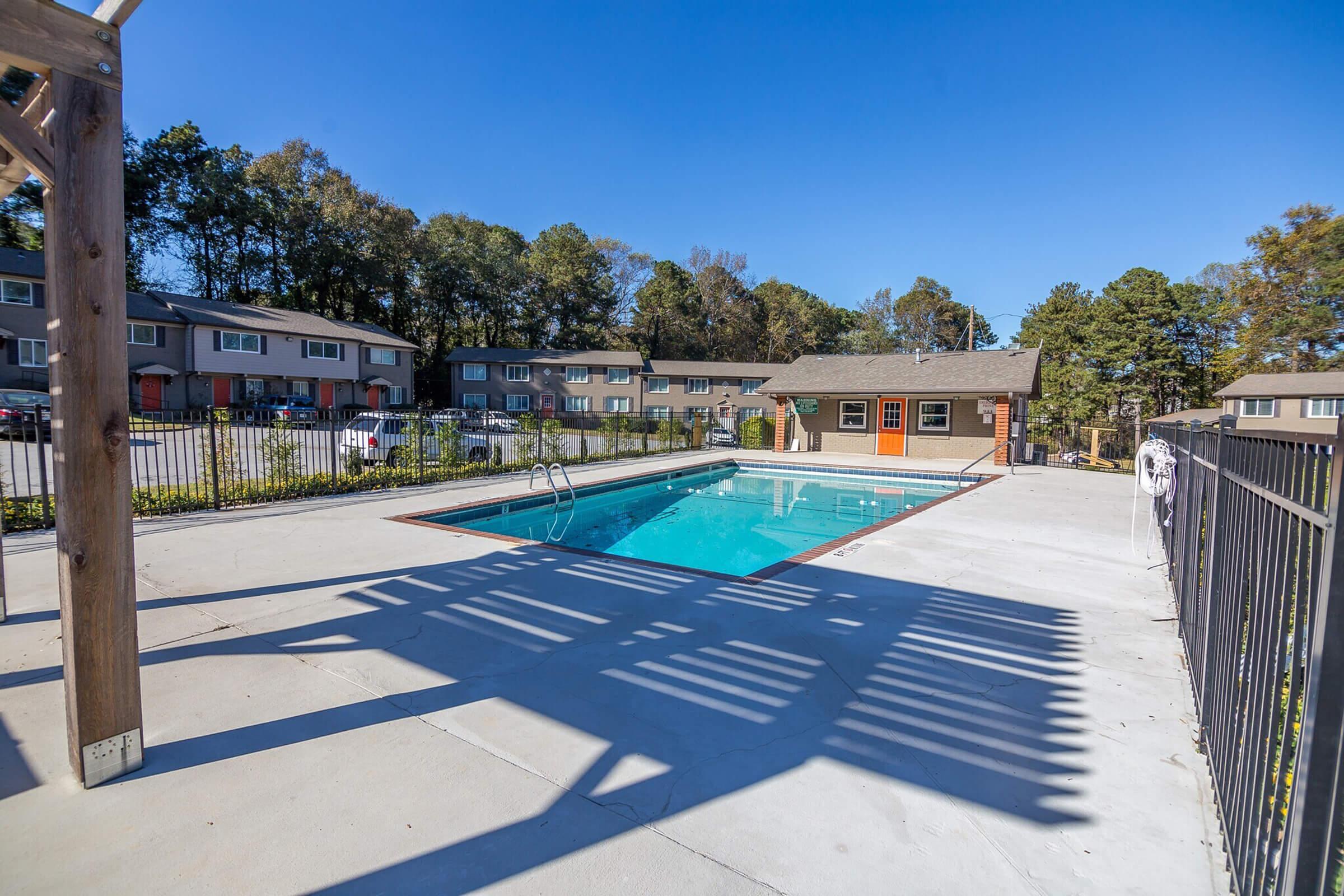 A clean outdoor swimming pool surrounded by a fence, with a nearby building featuring large windows. The area is shaded by trees, and there are lounge chairs alongside the pool, creating an inviting atmosphere for relaxation and leisure. Clear blue sky overhead.