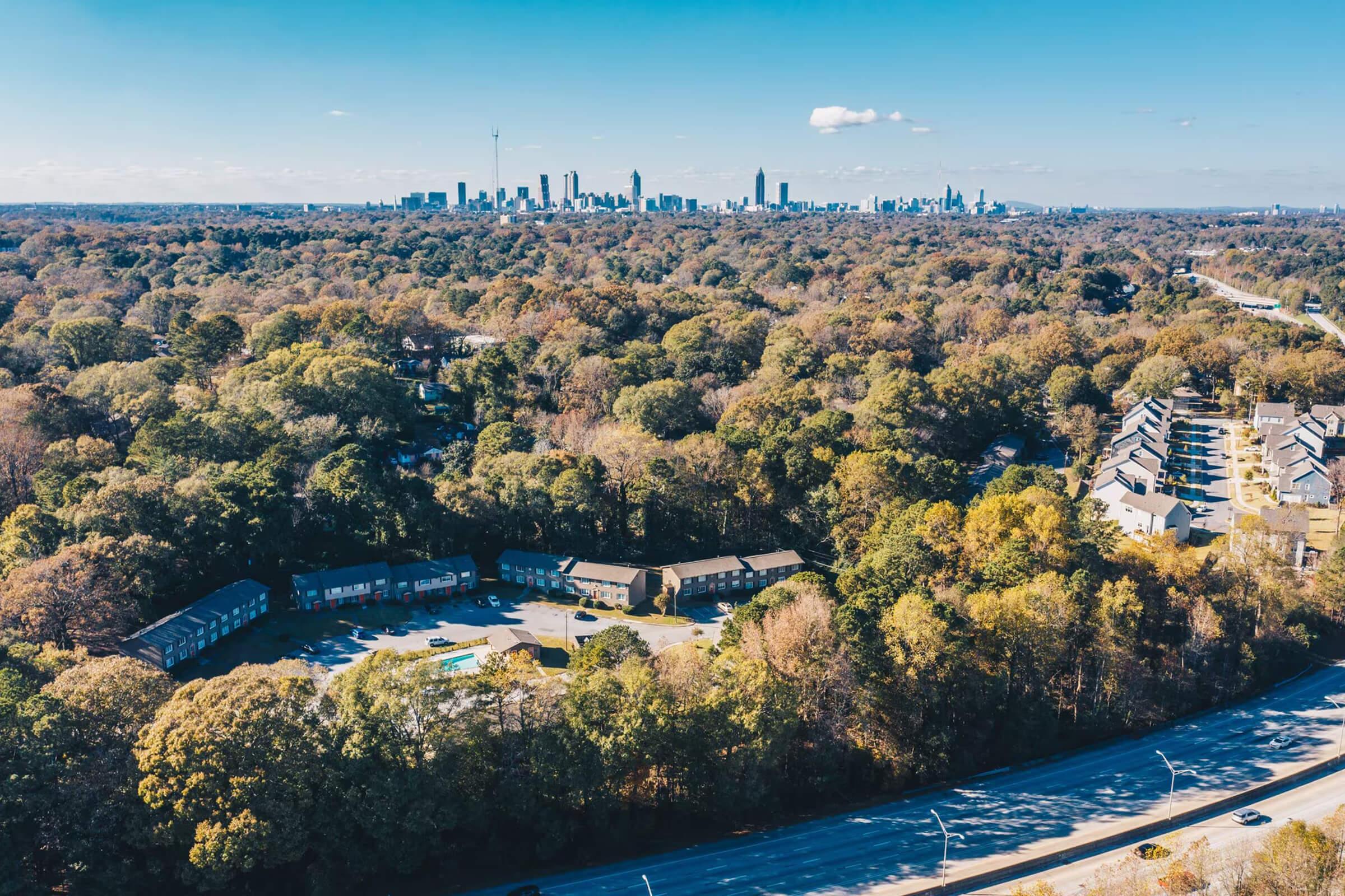 Aerial view of a city skyline in the distance, surrounded by lush green and autumn-colored trees. In the foreground, there are low-rise residential buildings and a road, with clear blue skies overhead and a few clouds above the skyline.
