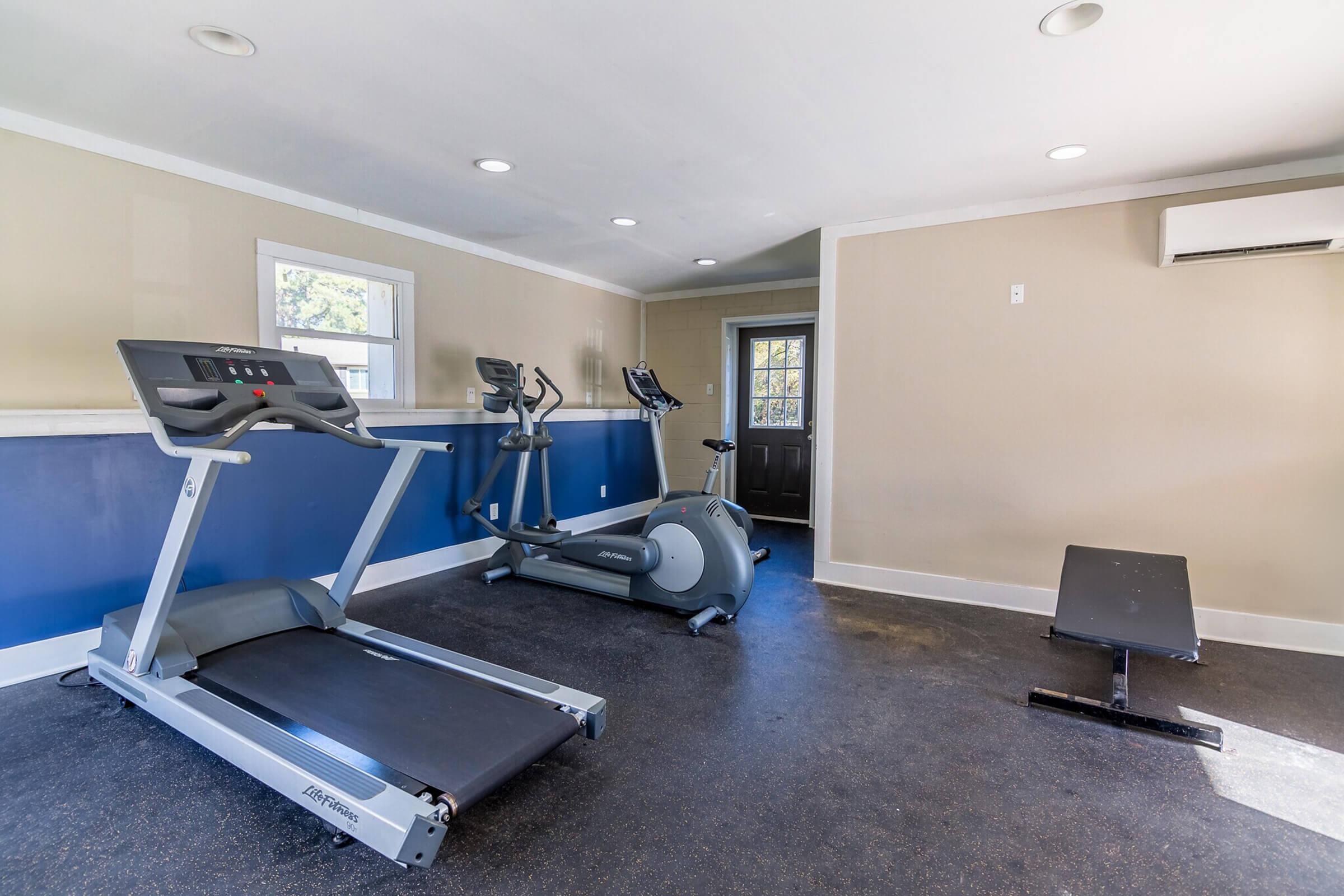 A small, bright fitness room featuring exercise equipment, including a treadmill, elliptical machine, and a weight bench. The walls are painted beige and blue, with ample natural light coming from a window. A door leads outside, and the floor is finished with dark rubber.