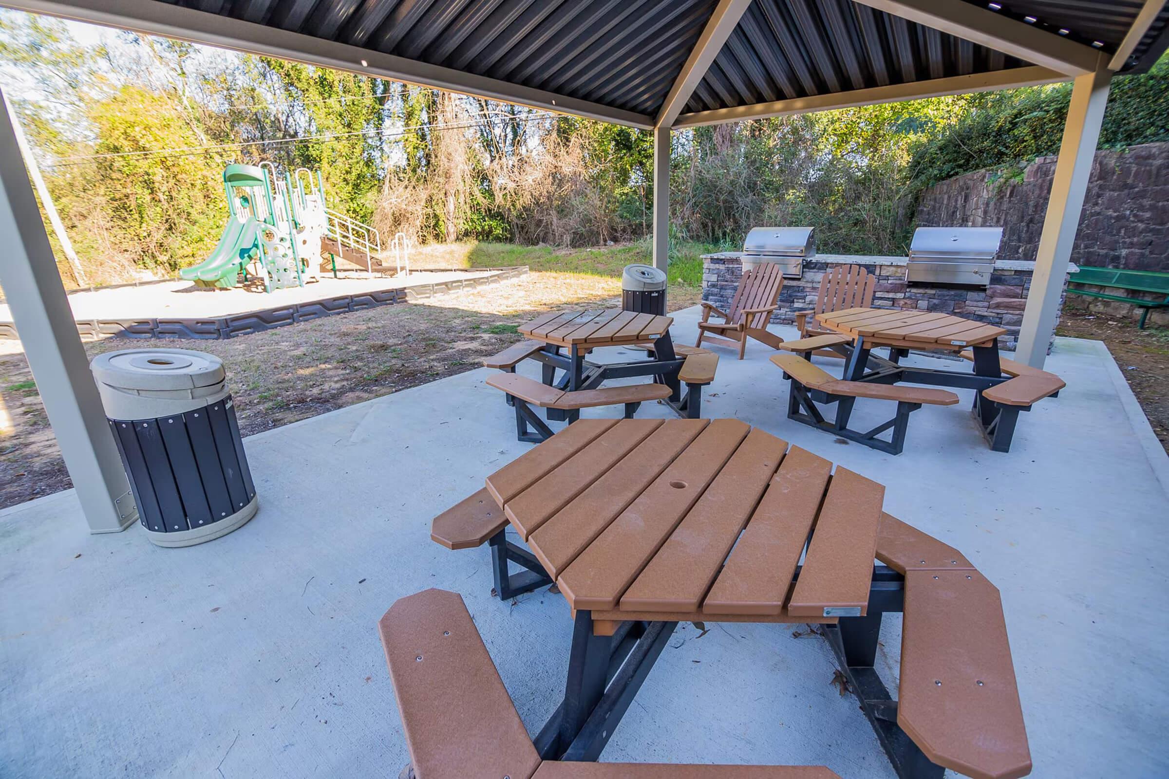 A picnic area with multiple octagonal tables under a roof, featuring park grills in the background. A green playground structure is visible to the side, with a trash can nearby and trees lining the background, creating a serene outdoor space.
